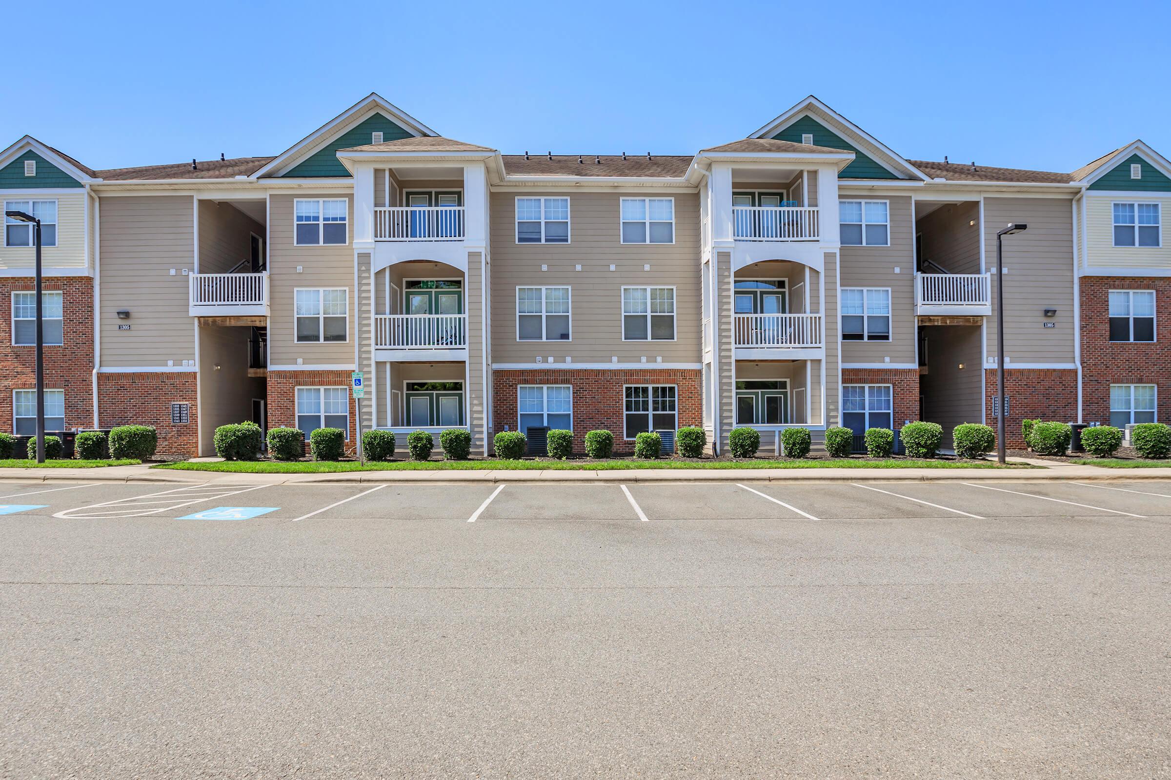 A modern multi-story apartment building with a mix of tan and brick exterior. The building features multiple balconies, large windows, and well-maintained landscaping. A parking lot is visible in front with marked parking spaces. The sky is clear and blue.