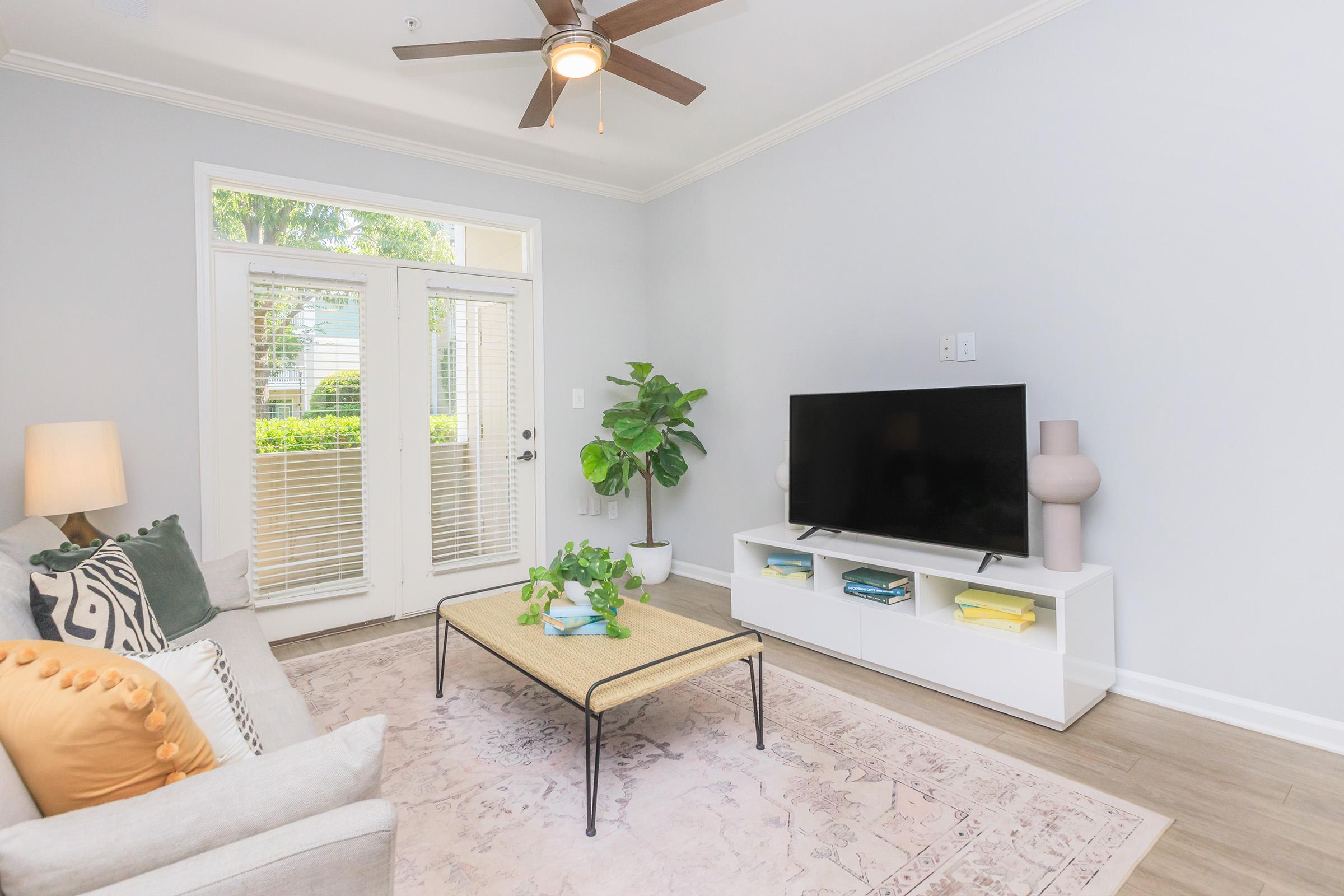 A cozy living room featuring a light-colored sofa with decorative pillows, a coffee table with a plant centerpiece, and a modern TV stand with neatly arranged items. Large windows provide natural light, and a ceiling fan adds a touch of comfort. The walls are painted a soft gray, creating a welcoming atmosphere.