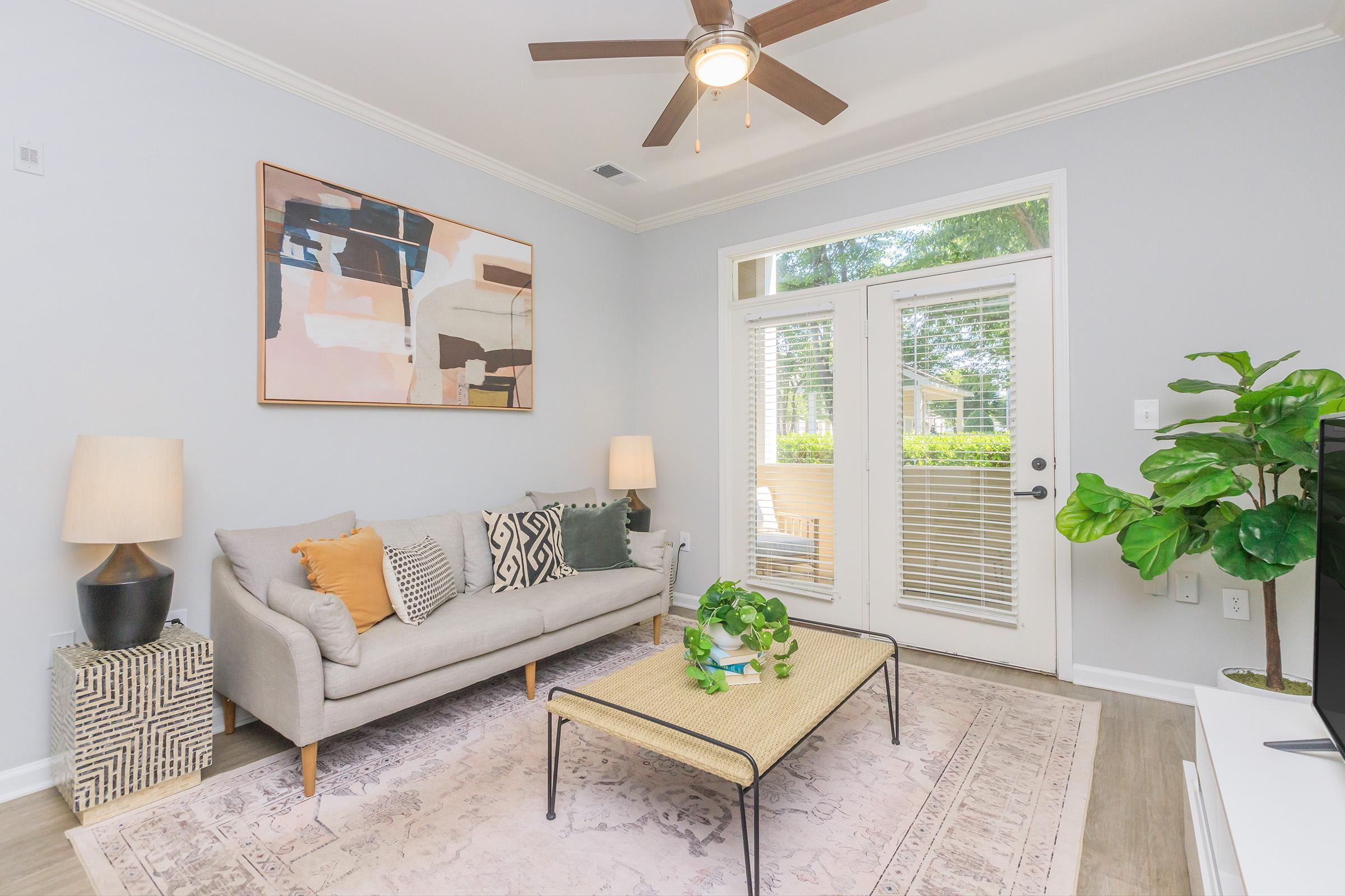 A bright and modern living room featuring a light grey sofa with decorative pillows, a coffee table with a plant centerpiece, and a large window letting in natural light. Two stylish lamps are positioned beside the sofa, and a colorful abstract painting hangs on the wall. A green plant adds freshness to the space.