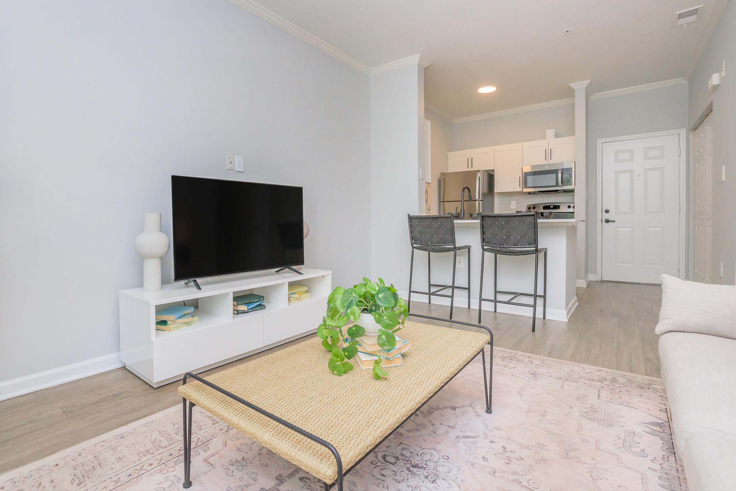 A modern living room featuring a flat-screen TV on a white media console, a decorative coffee table with a plant, and a light-colored sofa. The space has hardwood floors and leads to a kitchen area with bar stools, creating an open and inviting atmosphere.