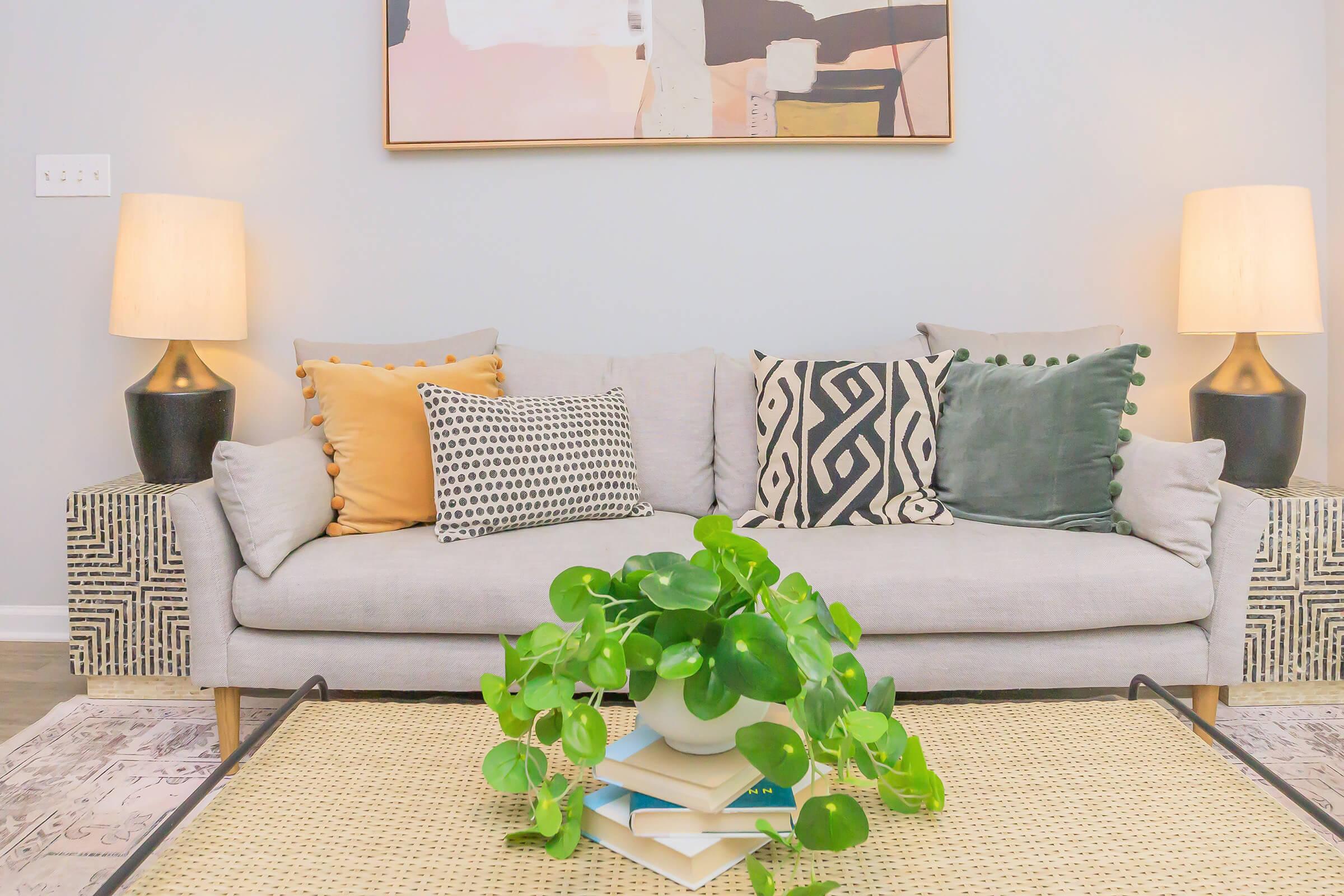 A modern living room featuring a light gray sofa adorned with decorative pillows in various patterns and colors. Two stylish lamps provide ambient lighting, and a coffee table displays a stack of books topped with a small green plant in a white pot, creating a cozy atmosphere.
