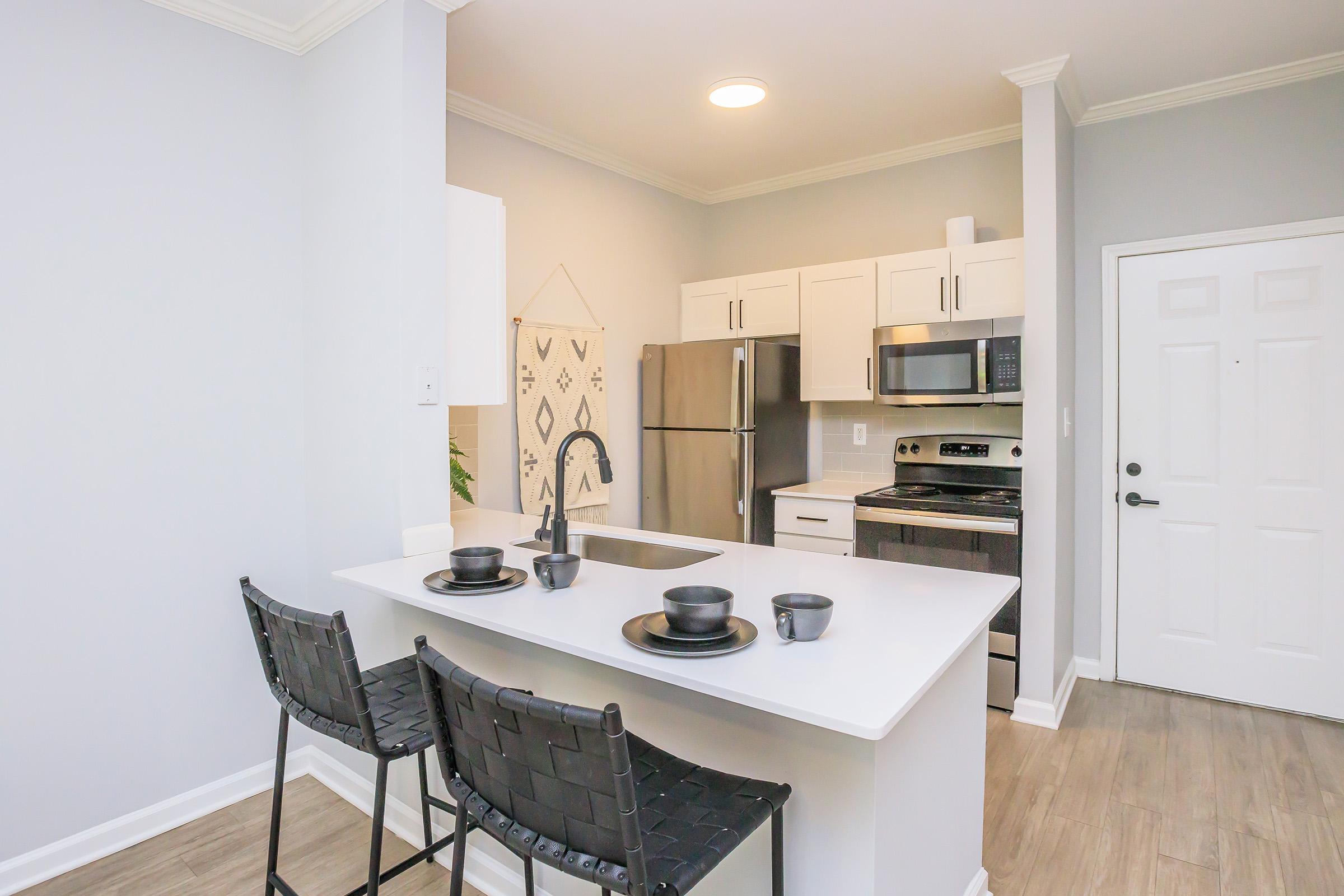 Modern kitchen featuring a stainless steel refrigerator, stove, and microwave. There is a white kitchen island with two black woven bar stools. The decor is minimalistic, with light gray walls and a door leading outside. Simple dishware is set on the island.