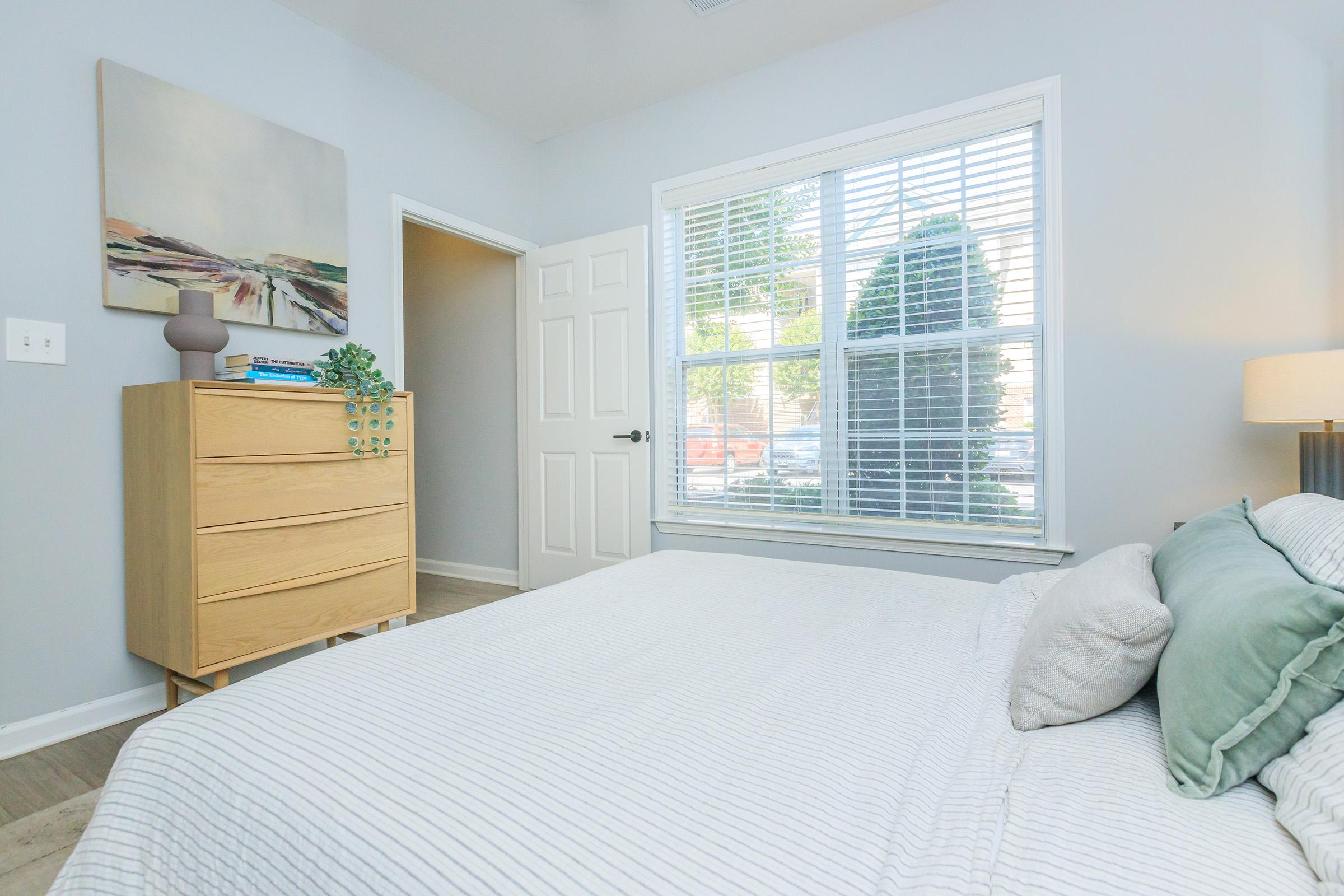 A bright and airy bedroom featuring a light-colored bed with cozy pillows, a wooden dresser against the wall, and a large window allowing natural light to pour in. A piece of abstract art hangs above the bed, and a small green plant decorates the dresser, adding a touch of greenery to the space.