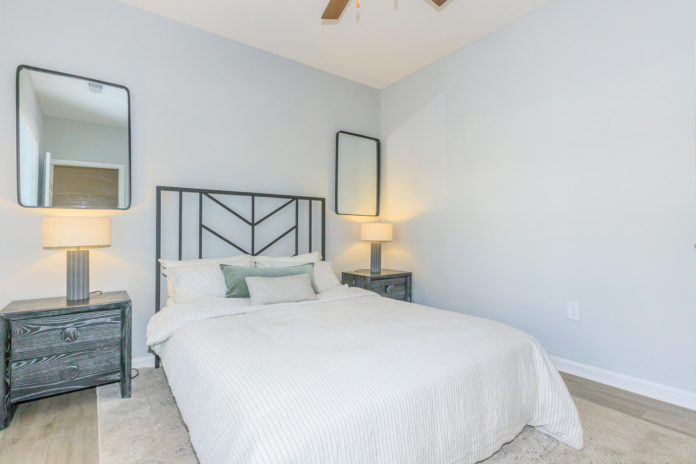 A cozy bedroom featuring a light-colored bed with decorative pillows, two modern bedside tables with lamps, and two wall mirrors. The room has a neutral color scheme, with a textured rug on the floor and a ceiling fan above, creating a serene and inviting atmosphere.