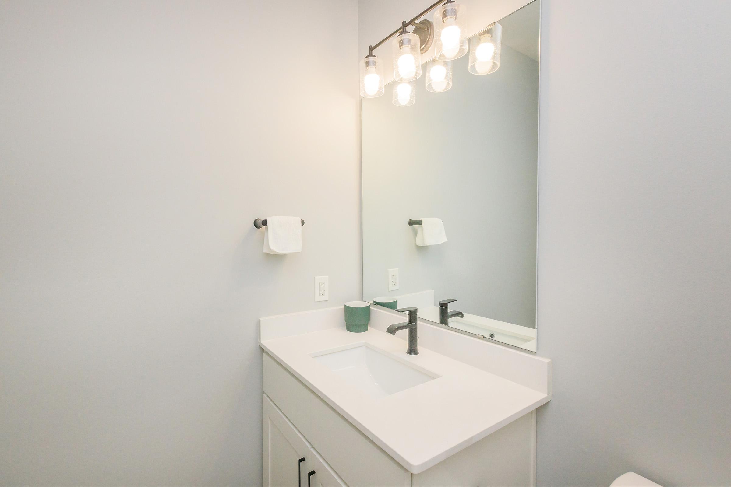 A modern bathroom with a white vanity featuring a rectangular sink, a sleek faucet, and a green cup. Above the sink, there is a large mirror flanked by a light fixture with three bulb-style lights. A white hand towel is hung on a towel bar beside the sink, against a light gray wall.