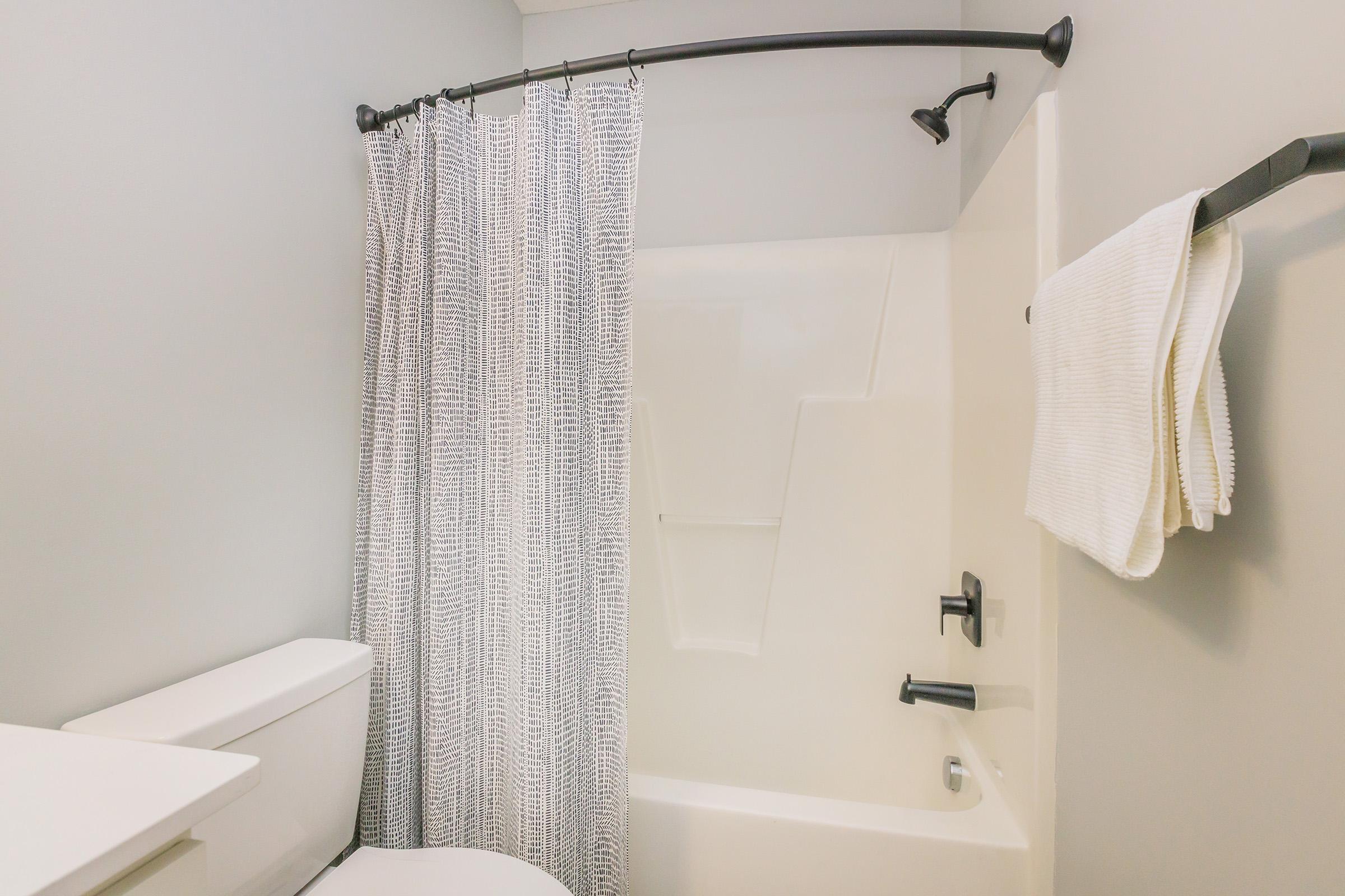 A clean and modern bathroom featuring a white bathtub with a gray and white striped shower curtain. There's a black towel rack with a neatly folded white towel, and a toilet positioned next to the bathtub. The walls are painted light gray, giving the space a bright and airy feel.