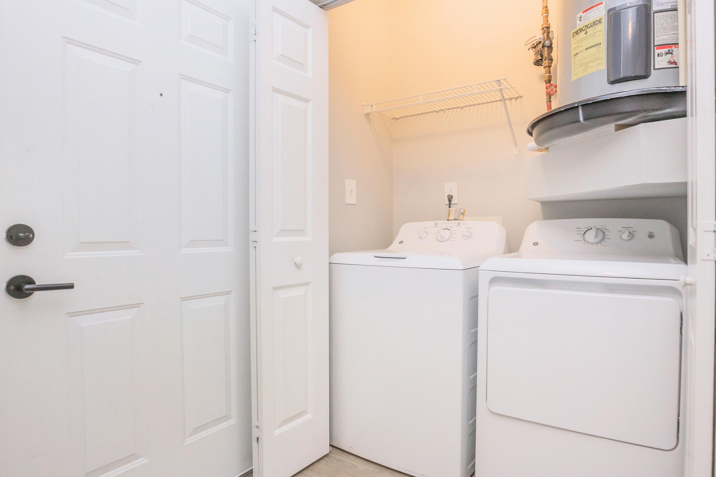 A small laundry room featuring a white washer and dryer side by side against a light-colored wall. There is a wire shelf above the washer, and a water heater is mounted on the wall. The room has a simple, clean design with a closed door visible on the left.