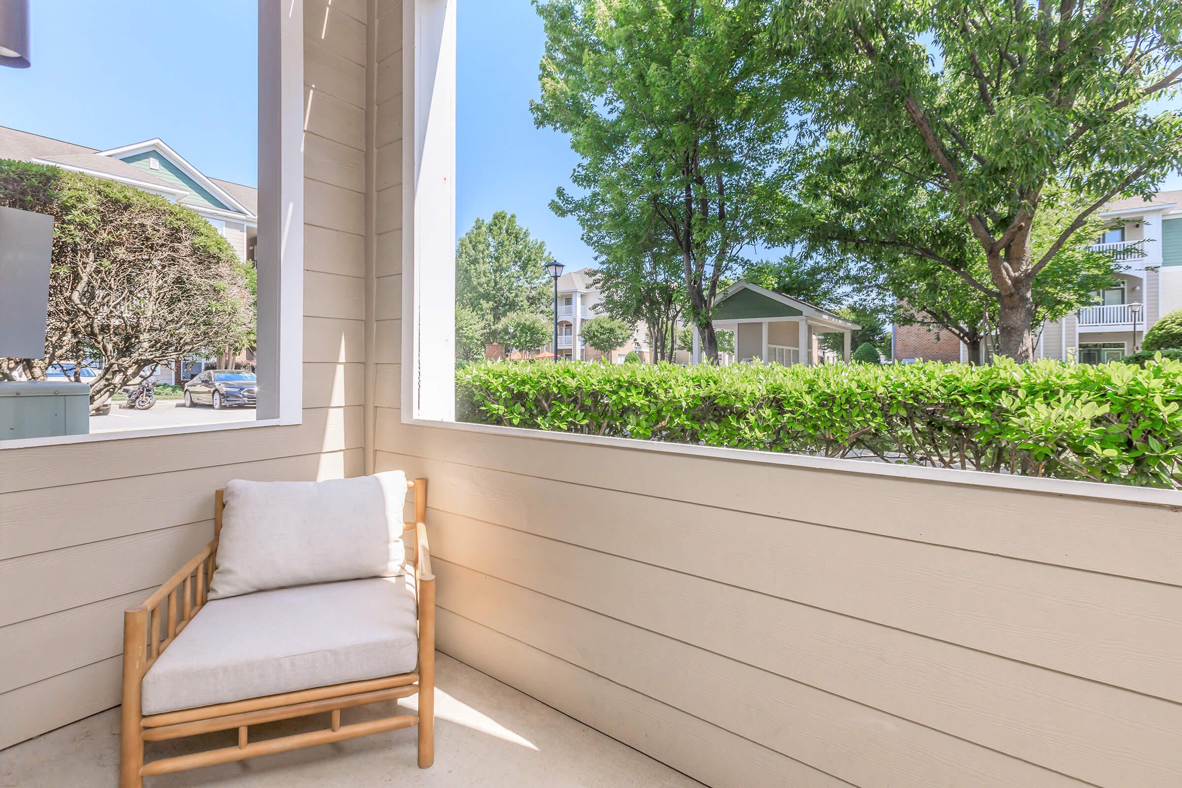 A cozy patio featuring a wooden chair with a soft cushion, surrounded by greenery. Bright sunlight illuminates the space, and a view of trees and nearby buildings can be seen through the open area.