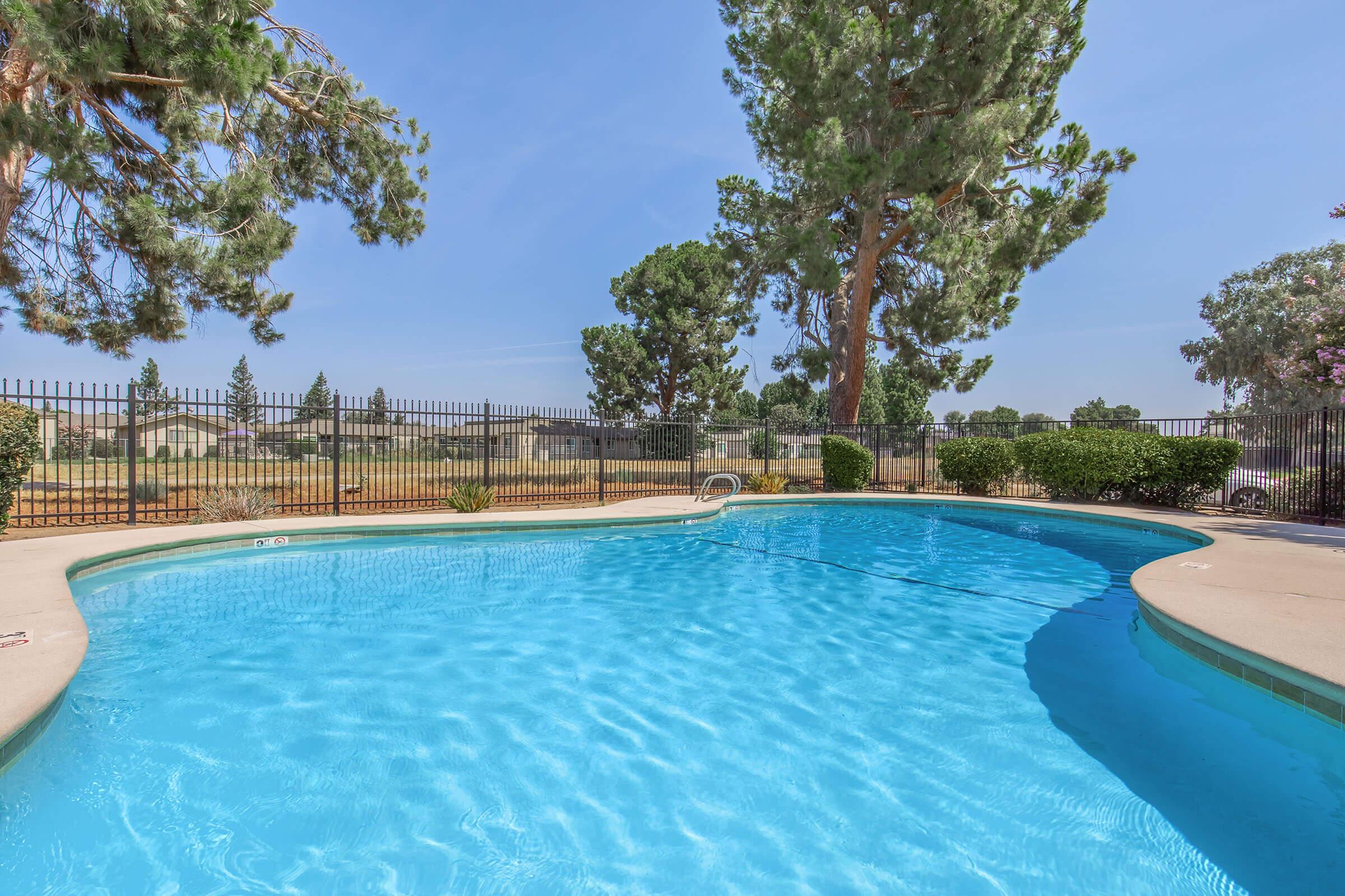 A clear blue swimming pool surrounded by a concrete deck, with tall trees in the background. A black fence encloses the pool area, and the sky is bright with a few wispy clouds. The scene conveys a tranquil, inviting atmosphere, ideal for relaxation or leisure activities.