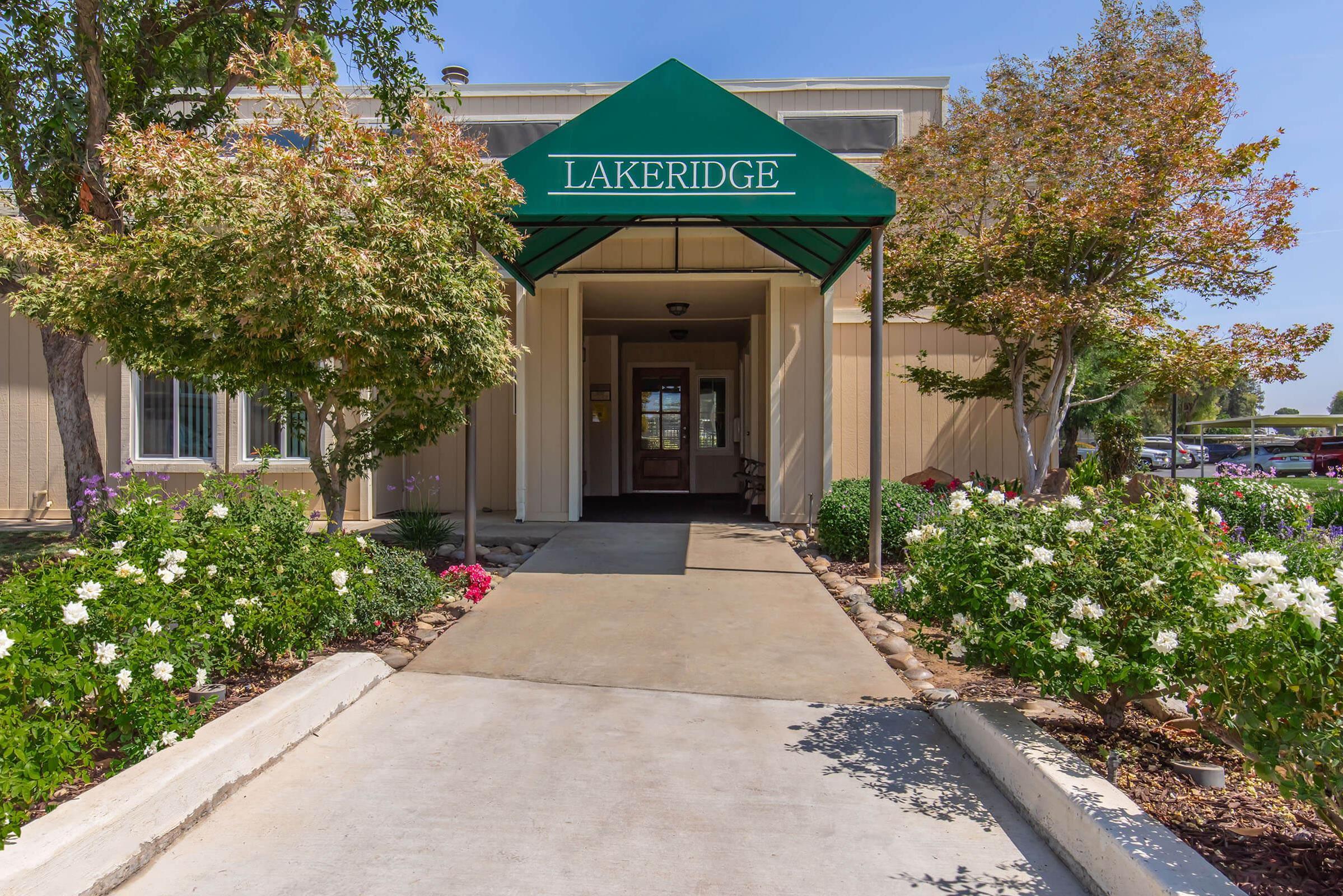 Entrance of a building with a green awning labeled "Lakeridge." A paved walkway leads to the door, flanked by flowering plants and trees. Clear blue sky in the background. The scene is well-lit and inviting, showcasing a clean and maintained exterior.