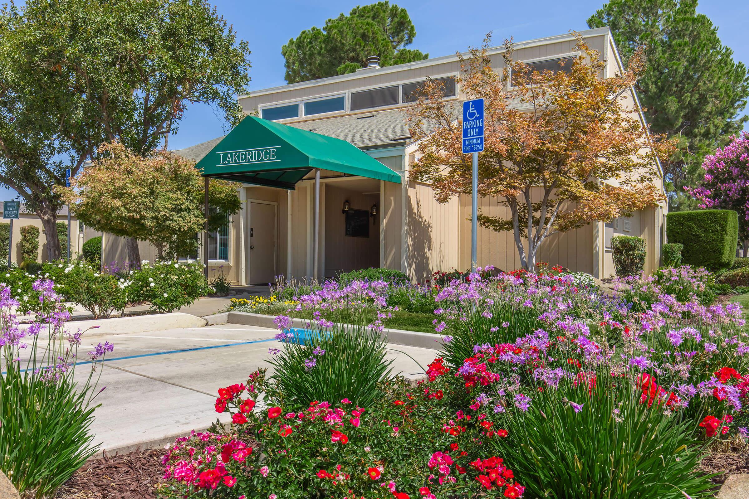 A charming single-story building with a green awning labeled "Lakeridge." Surrounding the entrance are colorful flower beds featuring red, pink, and purple blooms. Lush trees and shrubs provide greenery, and a blue handicap accessibility sign is visible nearby, set against a clear blue sky.