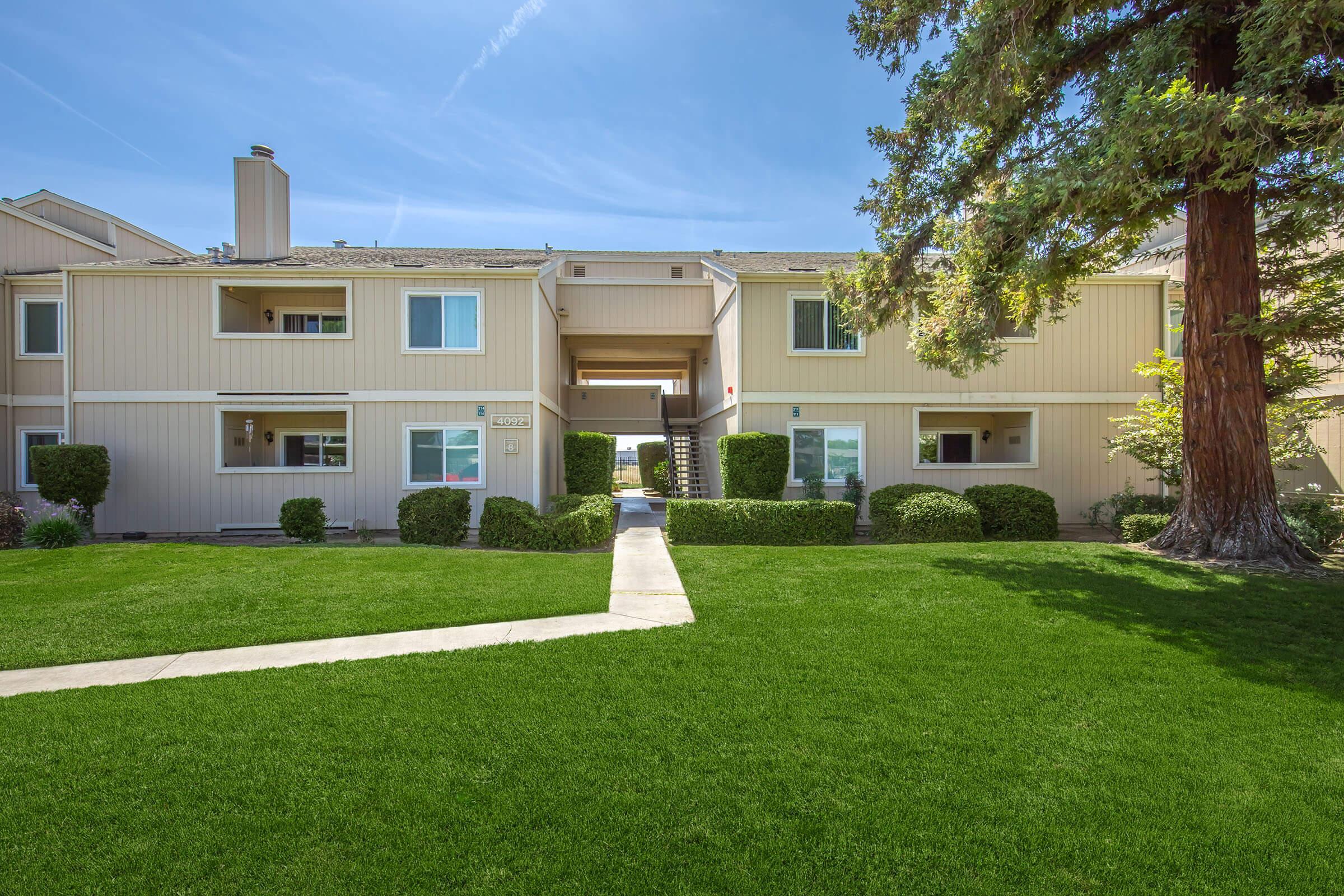 A well-maintained apartment complex with two-story buildings, featuring a green lawn and neatly trimmed bushes. Pathways connect the entrances, and a large tree provides shade. The sky is clear, indicating a sunny day.