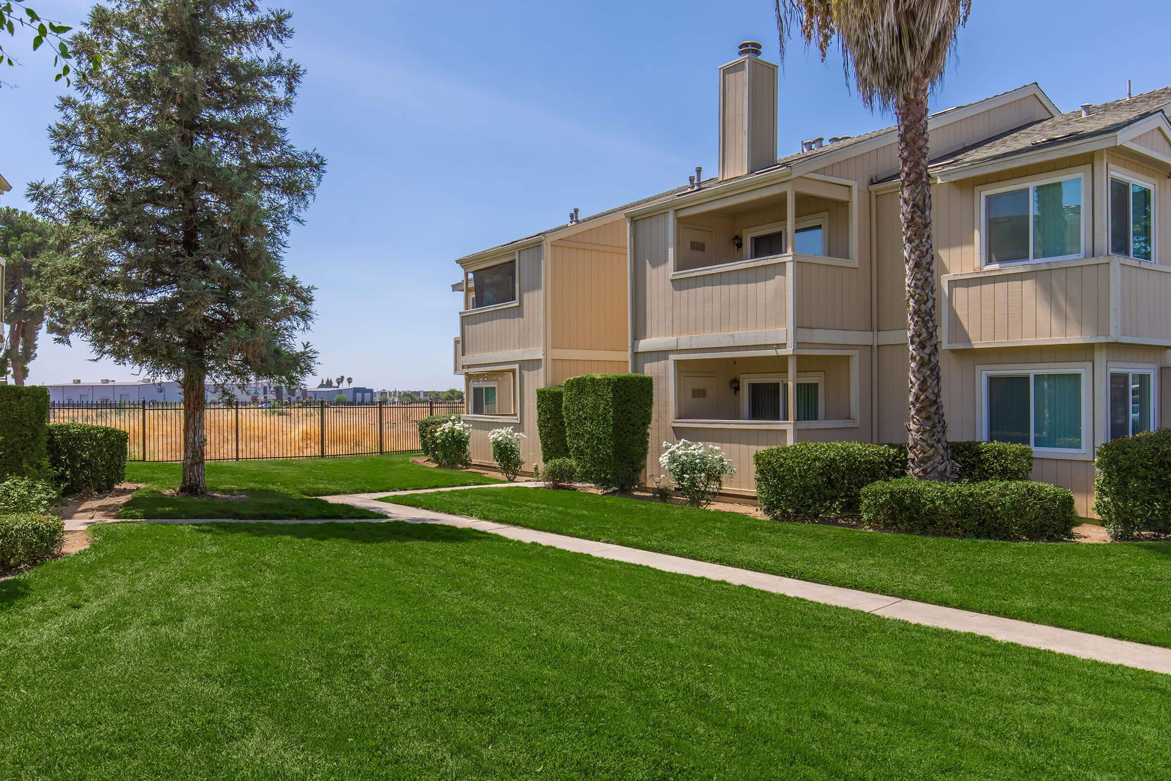 A well-maintained apartment complex featuring two buildings with balconies, surrounded by lush green lawns and neatly trimmed shrubs. In the background, a fenced area with open fields under a clear blue sky. Tall trees provide shade along the walkway leading to the entrance.
