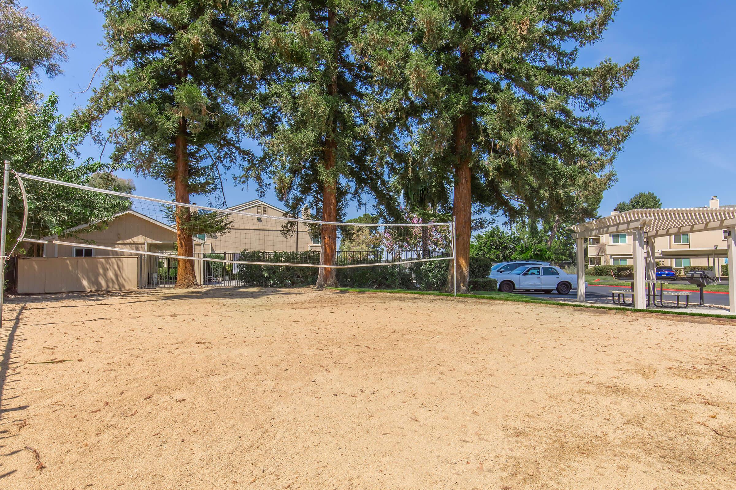 A sandy volleyball court surrounded by tall trees. In the background, there are residential buildings. A white car is parked nearby, and the sky is clear and blue. The area appears inviting for outdoor activities.