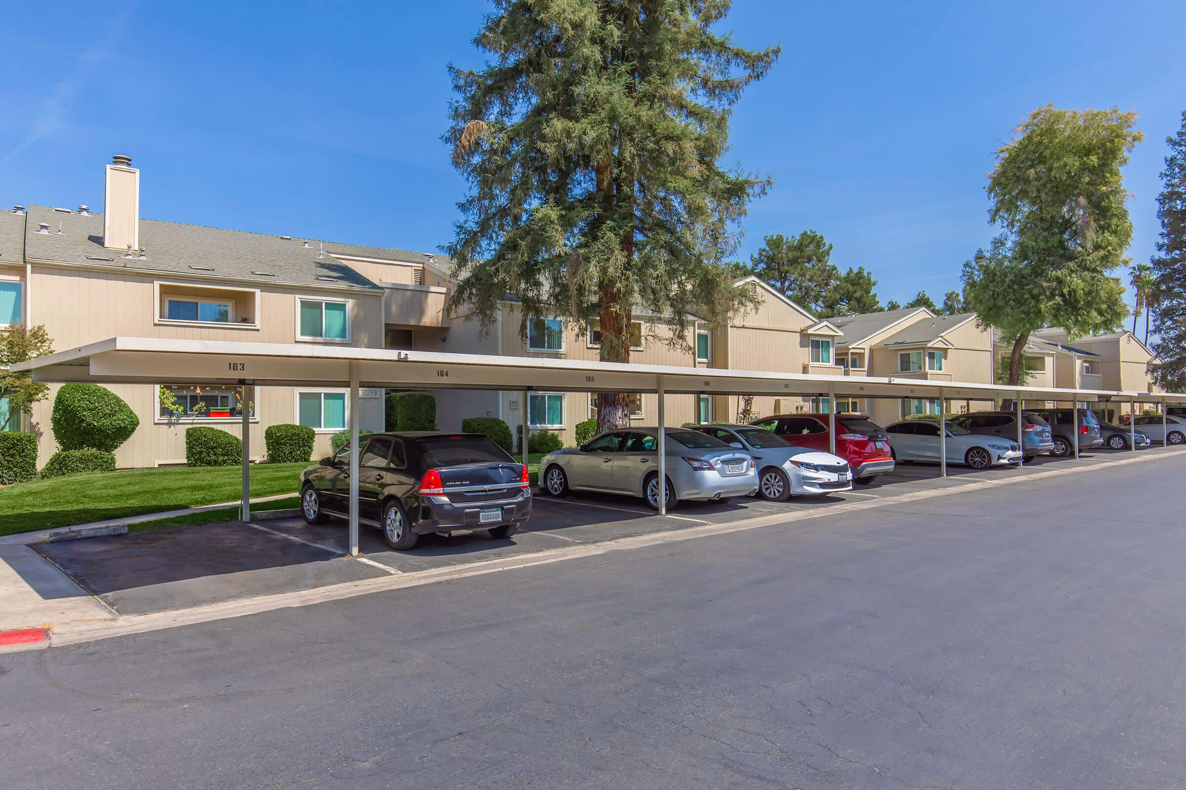 A row of covered parking spaces in front of a residential apartment complex. The complex features two-story buildings with a light beige exterior and green landscaping. Several parked cars are visible, and the scene is set under a clear blue sky.