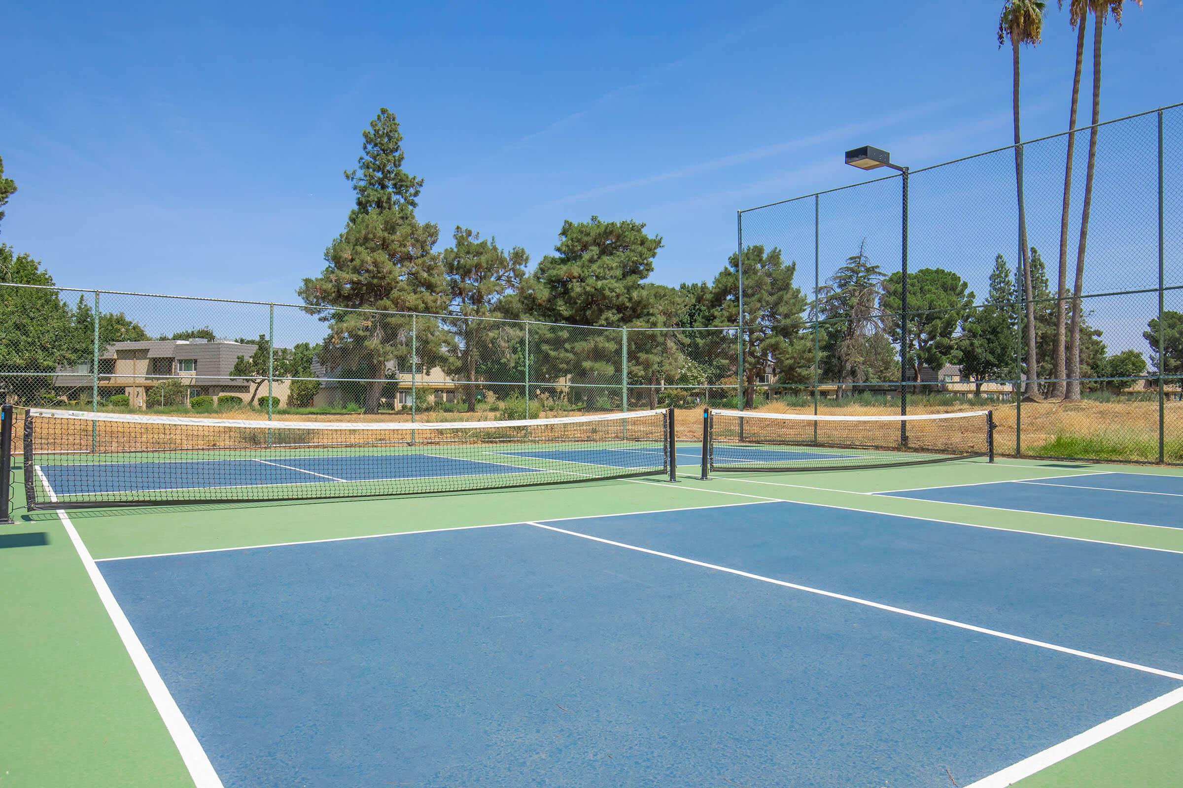 A view of tennis courts featuring blue and green surfaces, surrounded by trees and residential buildings in the background. The courts are well-maintained and under a clear blue sky, with a light post for evening play.