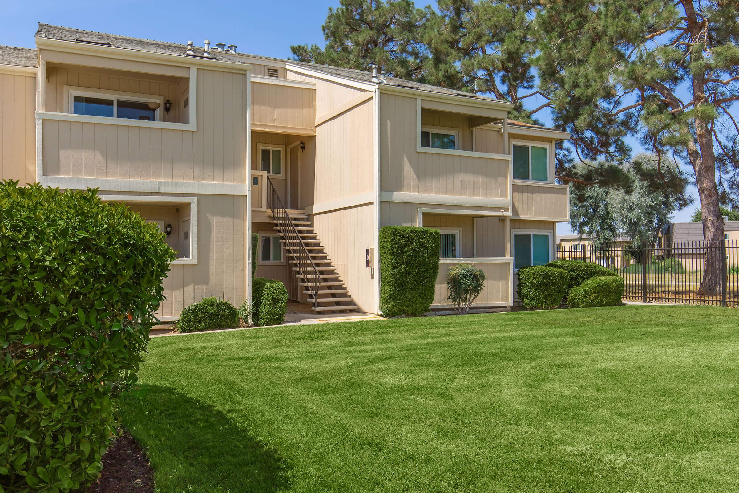 Exterior view of a two-story apartment building with a light-colored facade. The building features a stairway leading to the upper level and several windows. Surrounding the building is a well-manicured green lawn and neatly trimmed bushes, with trees in the background and a fenced area visible.