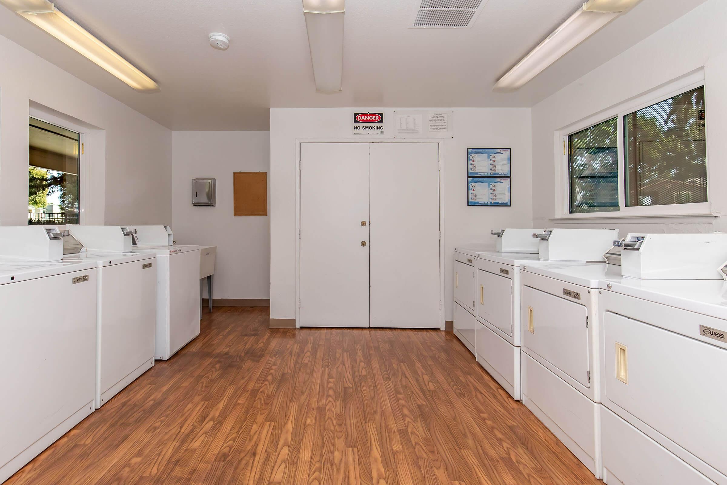 A clean laundry room featuring several white washing machines and dryers aligned against the walls. The space has light-colored walls and a wooden floor, with large windows allowing natural light. There is a sign that reads "No Smoking" on the wall, and various notices on blue papers are displayed nearby.