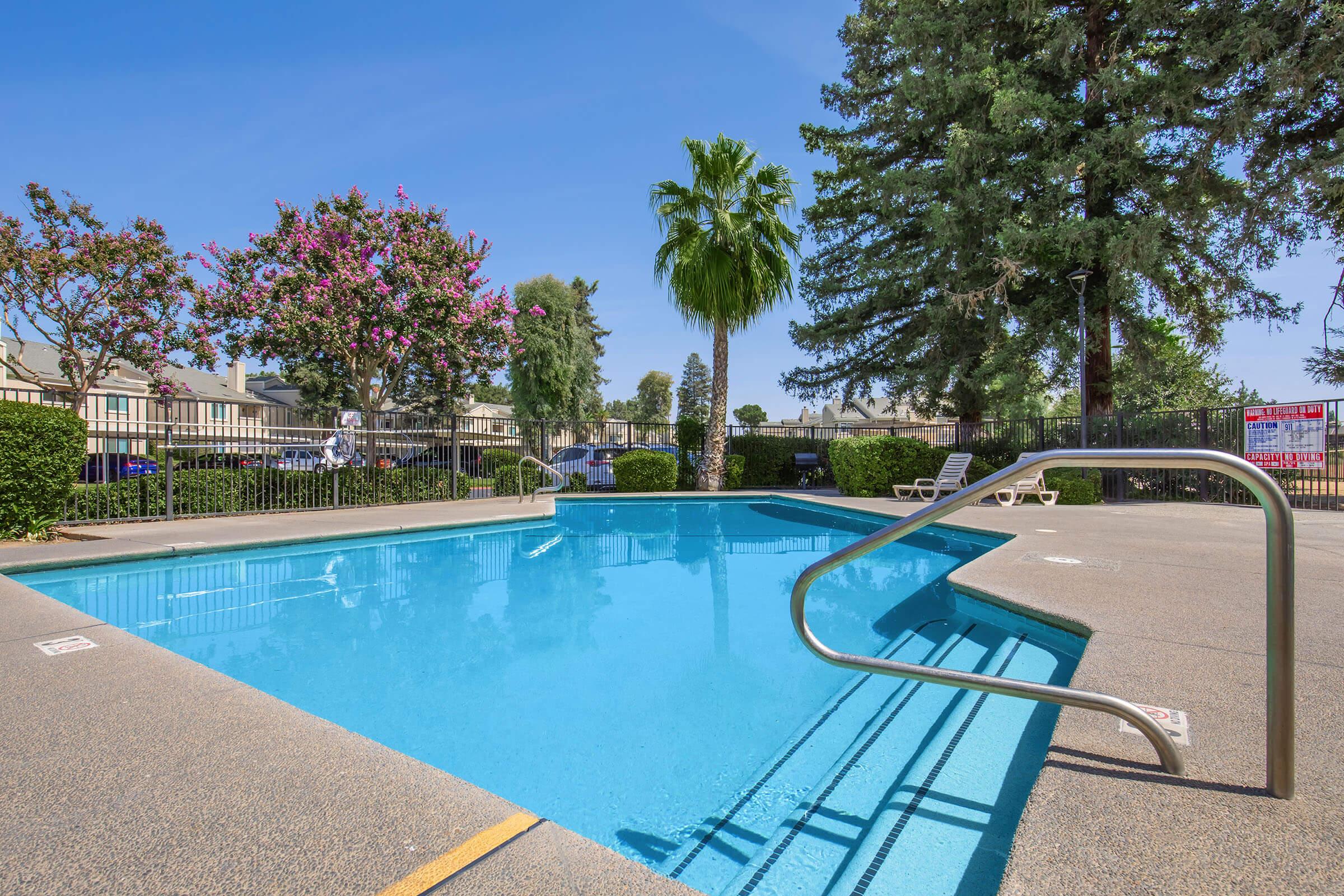 A clear blue swimming pool surrounded by palm trees and flowering plants. The pool area is enclosed with a fence, and lounge chairs are visible nearby. The sky is bright and sunny, enhancing the inviting atmosphere of the outdoor space.