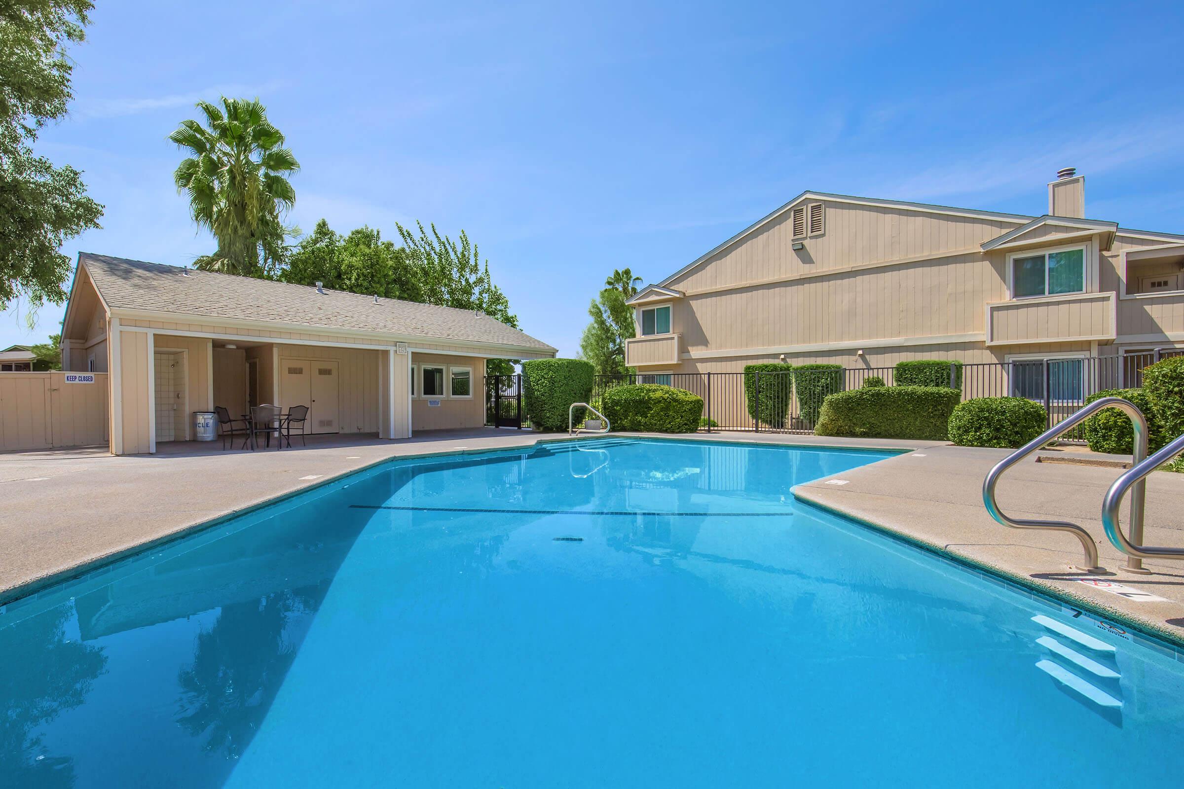 A clear blue swimming pool surrounded by greenery, with a small building nearby. The pool features a ladder and is bordered by well-maintained hedges, alongside an apartment complex in the background under a bright blue sky.