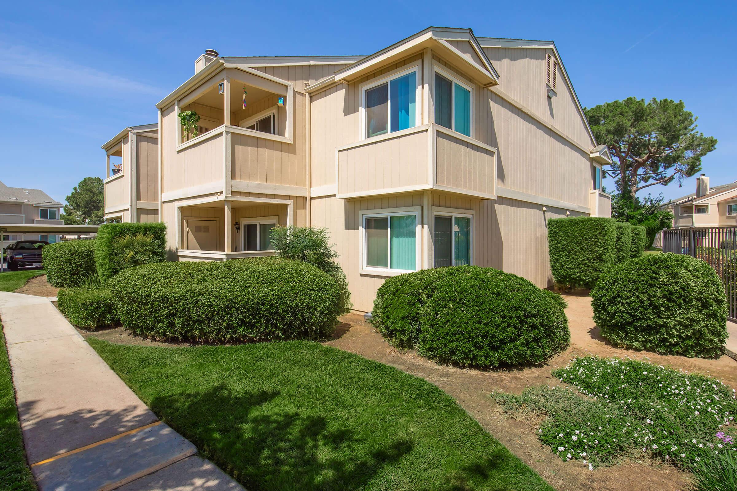 View of a modern two-story apartment building with beige siding and multiple windows. The building features small balconies, surrounded by neatly trimmed bushes and grass. A paved walkway leads to the entrance, with trees in the background and clear blue skies overhead.