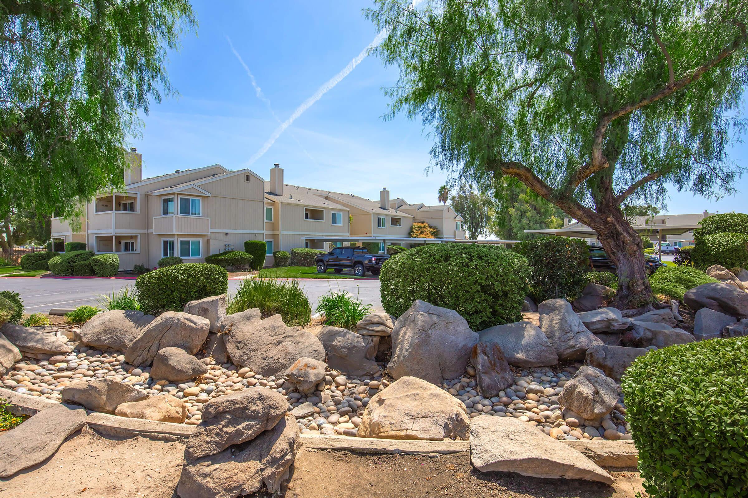 A landscaped area featuring large rocks, pebbles, and greenery. In the background, residential buildings are visible under a clear blue sky with a few clouds. Large trees with vibrant green foliage frame the scene, enhancing the outdoor atmosphere.