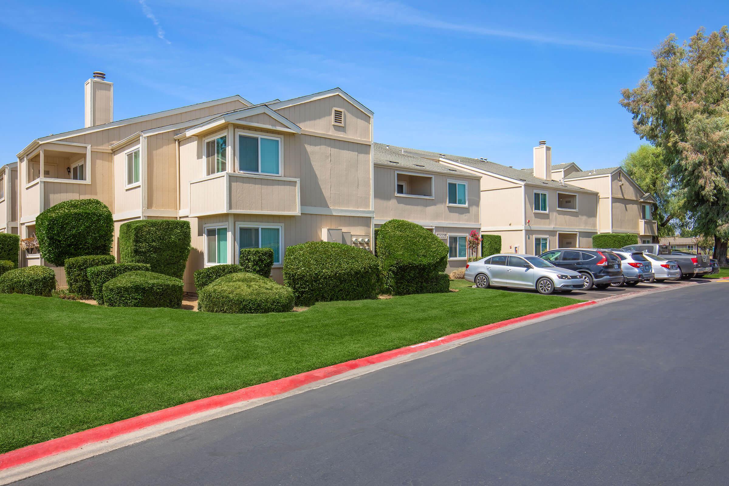 A view of a residential apartment complex featuring two-story buildings with light-colored exteriors, neatly trimmed hedges, and a well-maintained green lawn. Several cars are parked along the edge of a paved road. Clear blue skies enhance the pleasant atmosphere of the neighborhood.