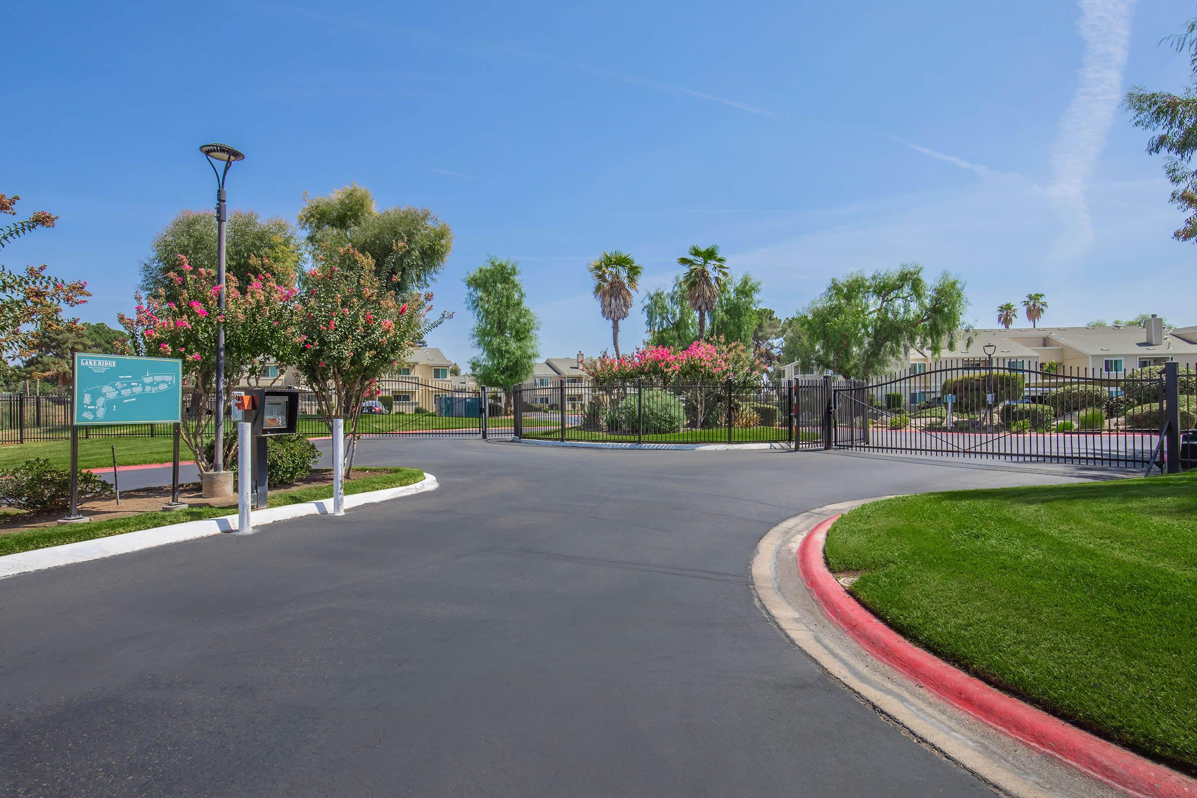 A gated entrance to a residential community, featuring flowering trees, palm trees, and well-maintained landscaping. The asphalt road leads to a secure entrance with a signboard visible on the left and a decorative gate ahead. Bright blue sky and greenery create a welcoming atmosphere.