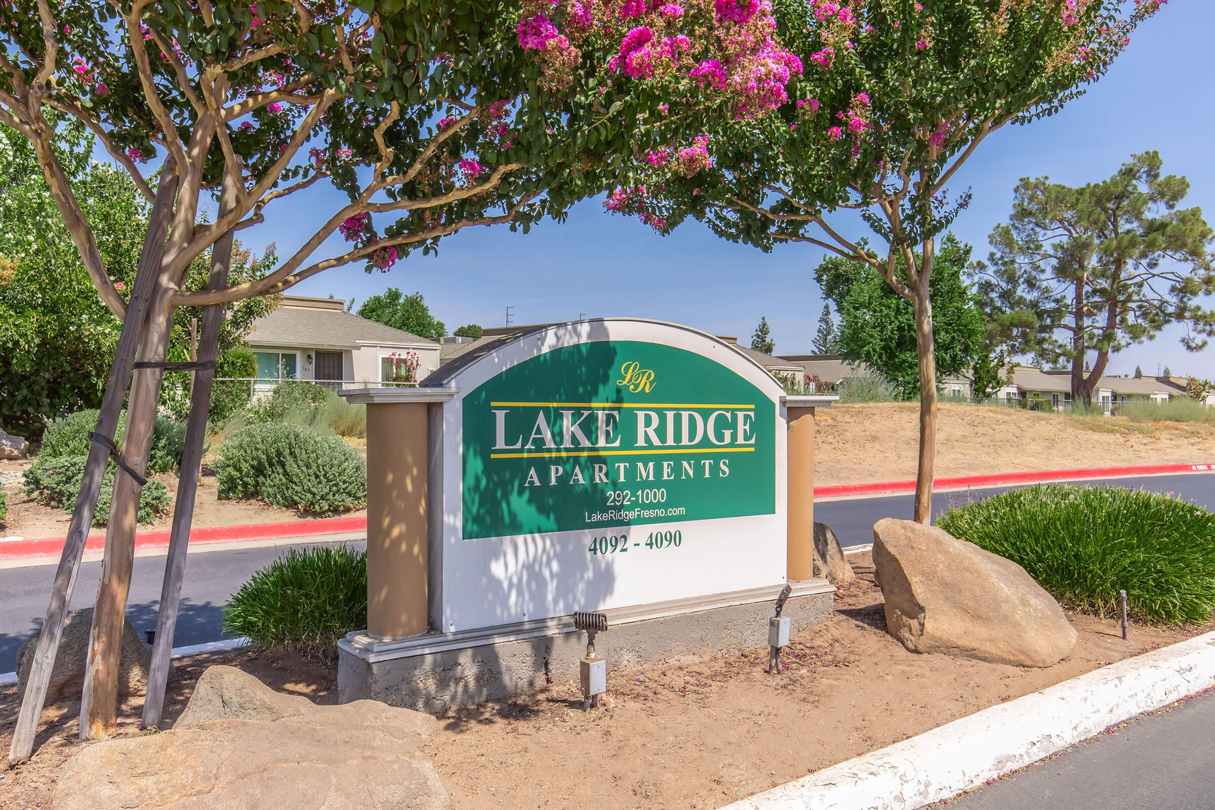 Sign for Lake Ridge Apartments, featuring the name "Lake Ridge" prominently displayed, along with the contact number 292-1000. The sign is surrounded by greenery and blooming flowers, with a clear blue sky in the background. The setting includes residential buildings visible beyond the sign.
