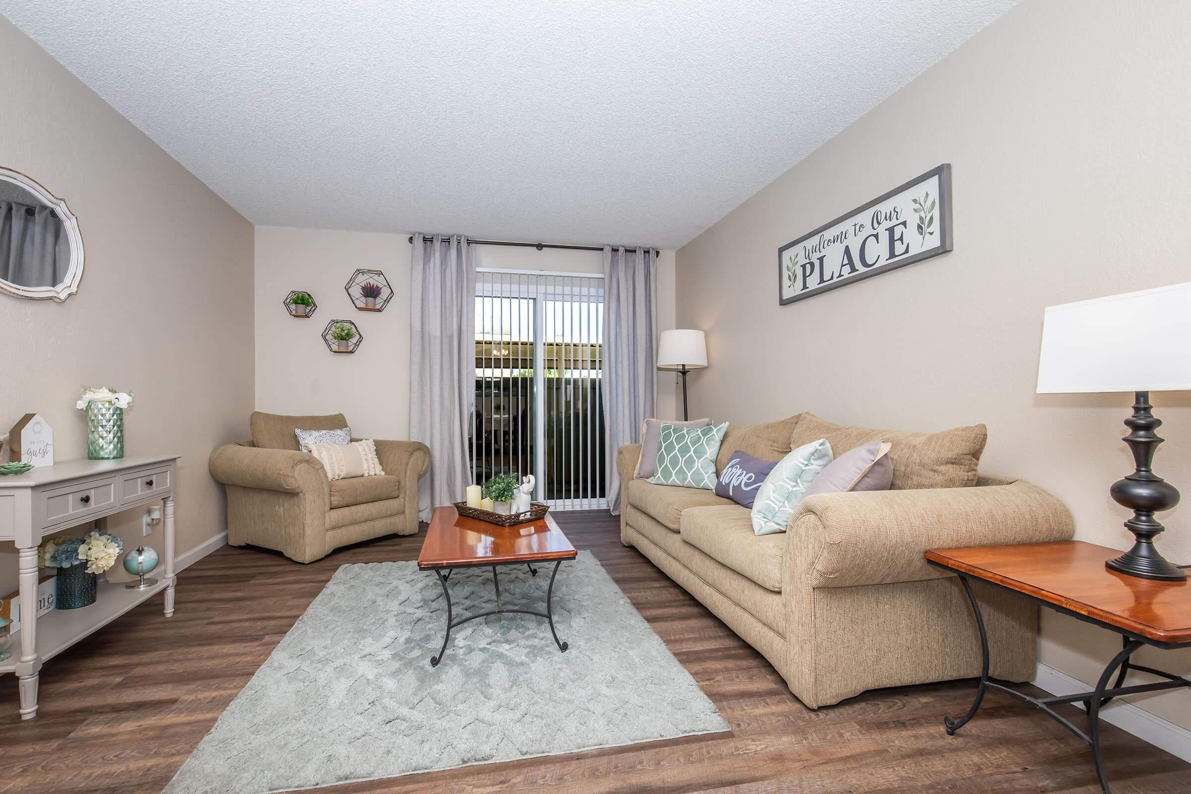 A cozy living room featuring two beige sofas adorned with decorative pillows, a wooden coffee table on a light rug, and a side table with a lamp. Wall decor includes round wall hangings, and a sliding door with curtains provides natural light and access to an outdoor space.