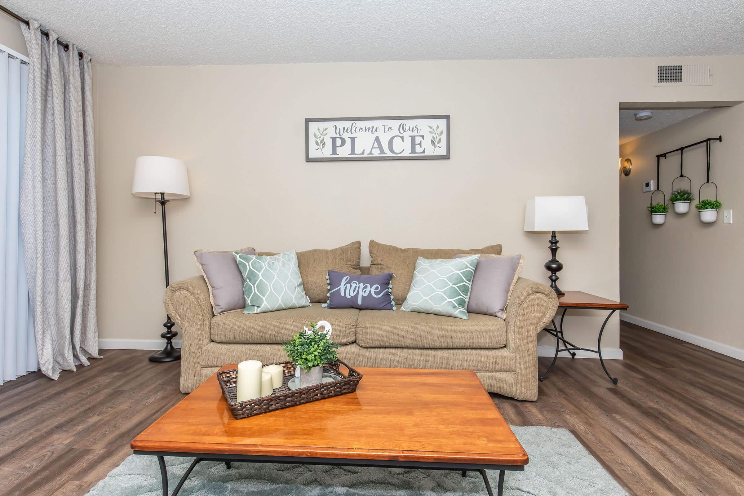 Cozy living room featuring a beige sofa with decorative pillows, a wooden coffee table with candles and a small plant, and a wall art piece that reads "Welcome to Our Place." Soft lighting from two lamps and neutral-colored walls create a warm and inviting atmosphere.