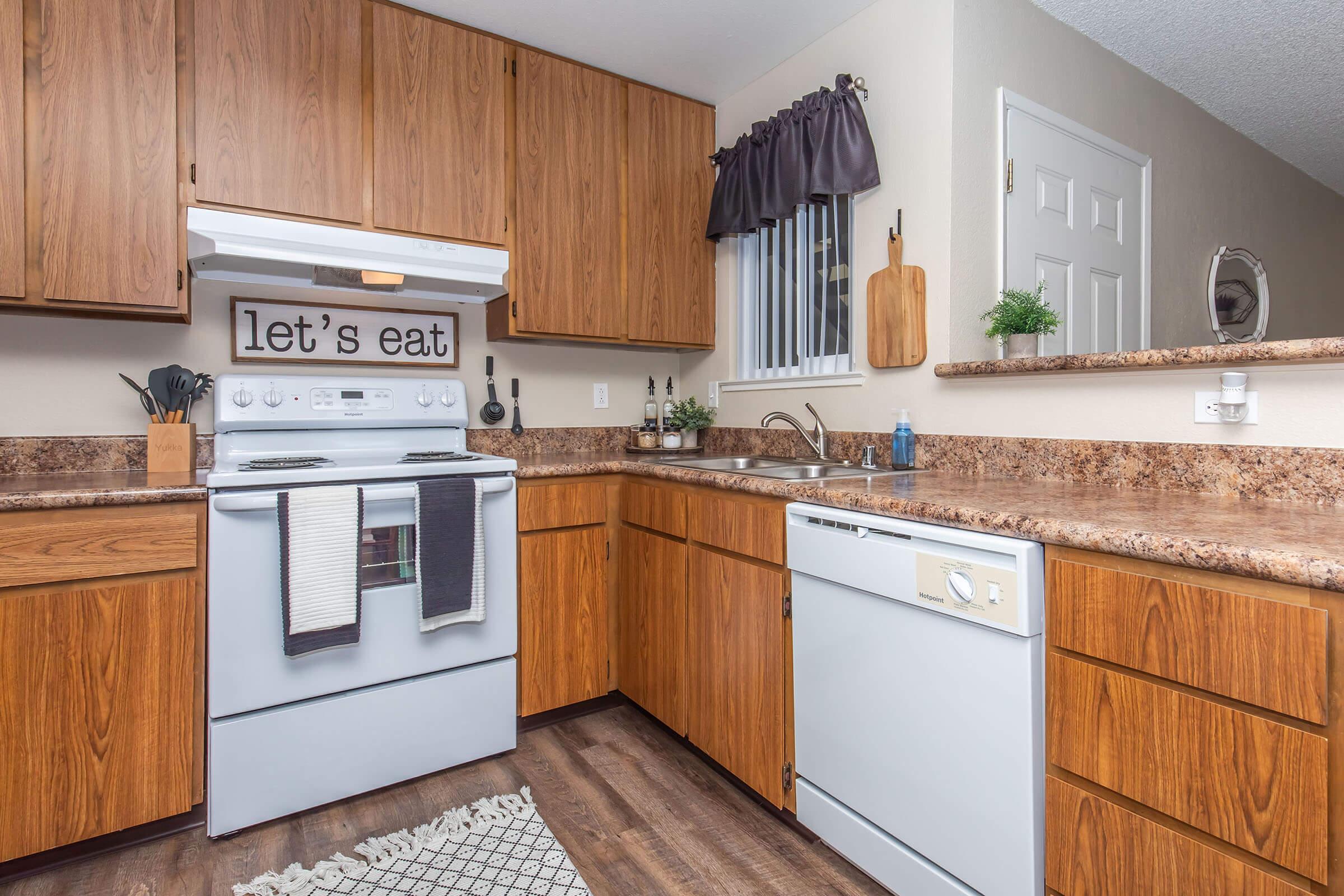 A modern kitchen featuring wooden cabinetry, a white stove and dishwasher, a stainless steel sink, and a countertop with brown granite. A sign that reads "let's eat" is displayed on the wall. The kitchen is well-lit with a window and has decorative plants and kitchen utensils visible.