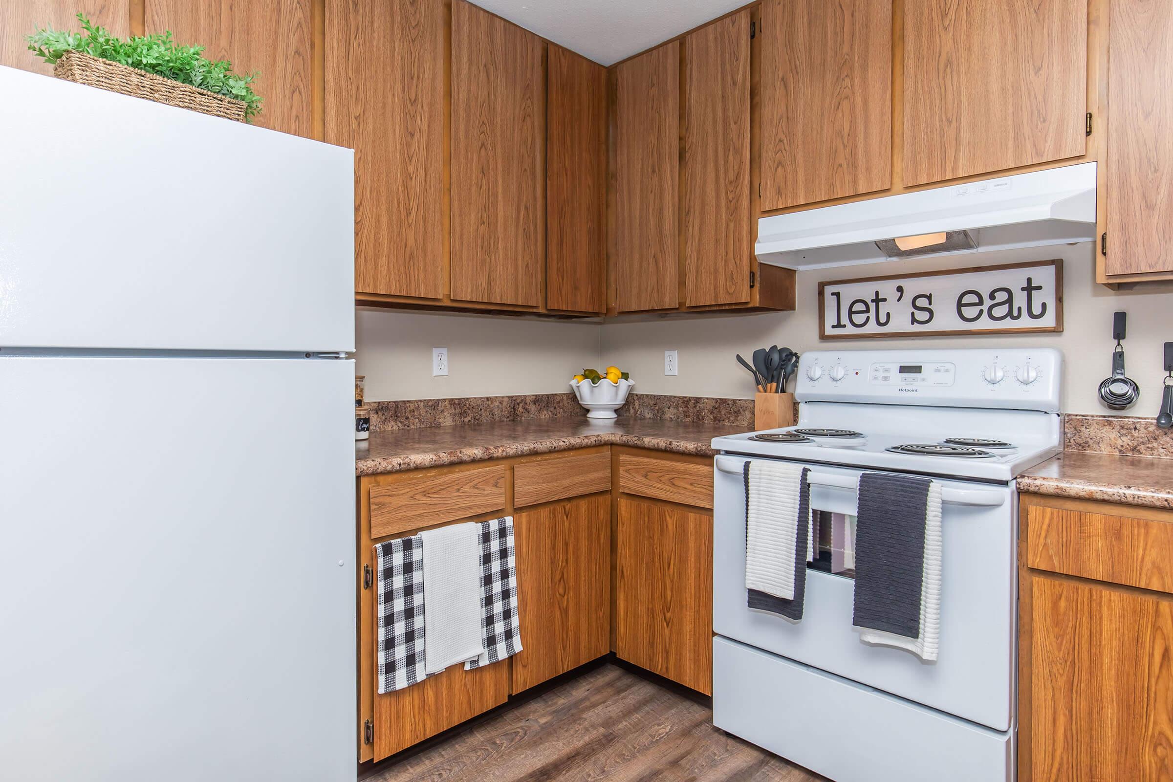 A cozy kitchen featuring wooden cabinets and a white refrigerator. The stove, beneath a range hood, is accompanied by hanging kitchen towels. A decorative sign on the wall reads "let's eat." A bowl of flowers is displayed on the countertop, adding a warm touch to the space.