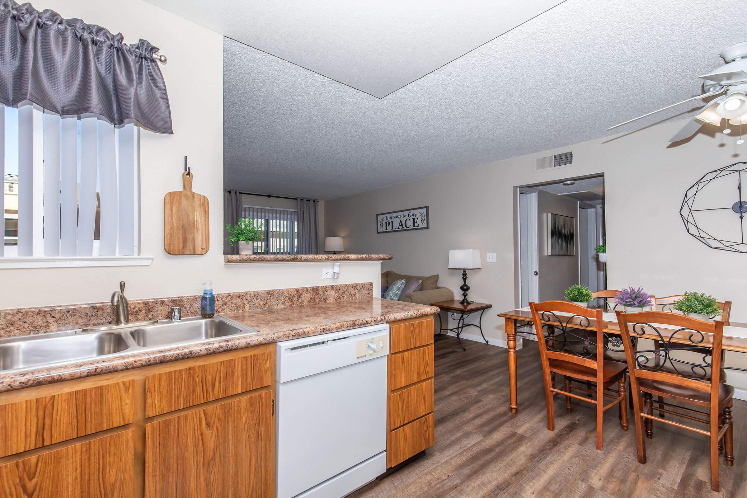 A modern kitchen with wooden cabinets and granite countertops, featuring a double sink and a dishwasher. In the background, there's a dining area with a wooden table and chairs, a ceiling fan, and decorative elements like plants and wall art. The space is well-lit and open, creating a welcoming atmosphere.