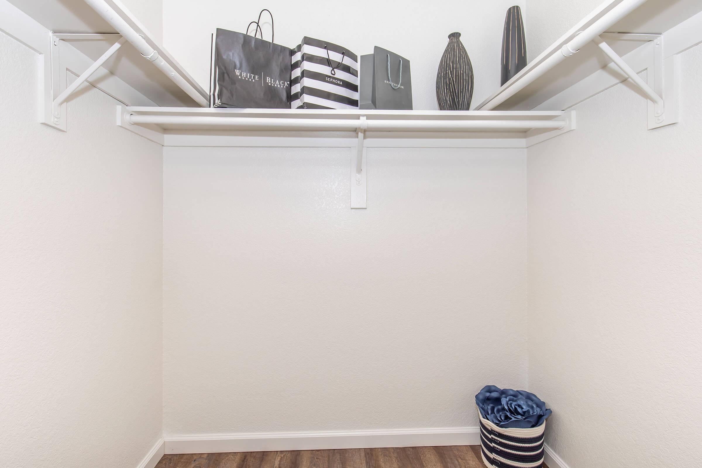 A neatly organized closet featuring a white shelving unit with shopping bags, a decorative vase, and a striped storage bin on the floor. The walls are painted a light color, and the flooring is a warm wood finish, creating a clean and modern aesthetic.