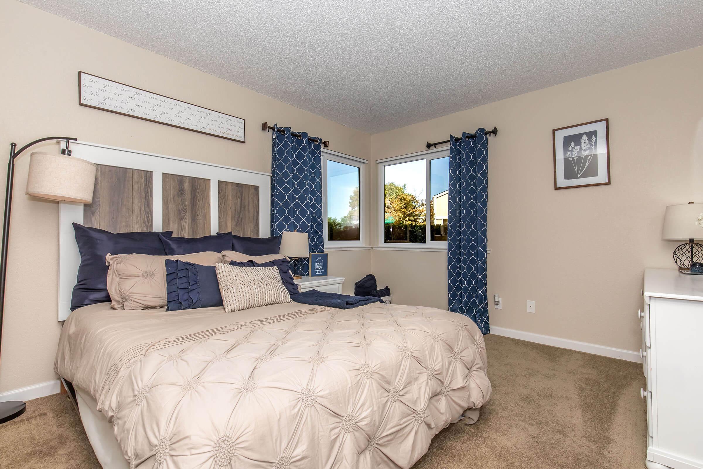 A cozy bedroom featuring a large bed with a beige tufted comforter and blue decorative pillows. There's a nightstand with a lamp beside the bed and two windows with blue curtains letting in natural light. A framed artwork hangs above the bed, and the room has neutral-colored walls and carpet.
