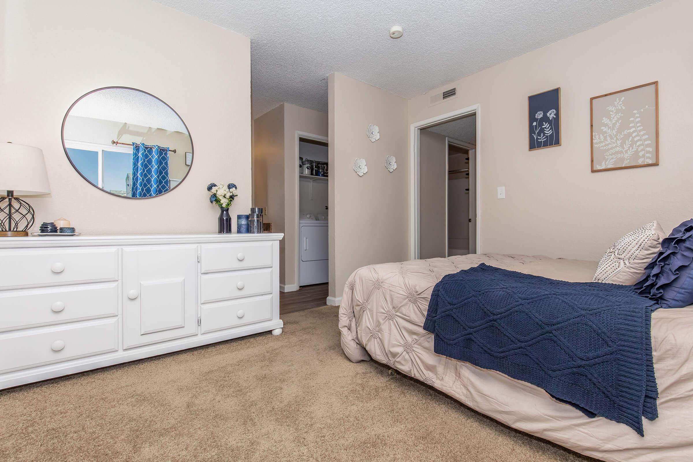 A modern bedroom featuring a comfortable bed with a beige quilt, a navy throw blanket, and decorative pillows. There is a white dresser with decorative items, a round mirror on the wall, and framed artwork. A closet door can be seen in the background, providing additional storage. The room has soft lighting and a warm color palette.