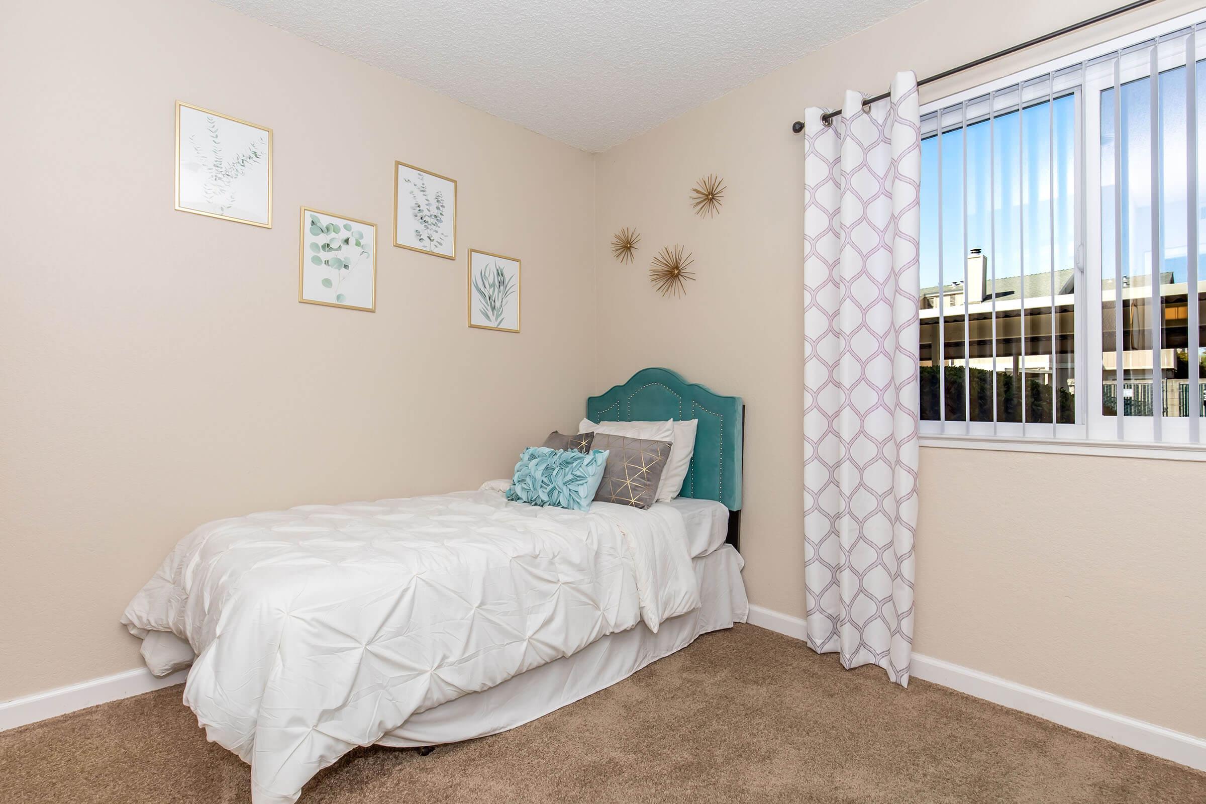 A cozy bedroom featuring a single bed with white bedding and decorative pillows. The walls are adorned with framed botanical art, and a window with vertical blinds allows natural light to enter. Soft beige carpet complements the serene color scheme, creating a tranquil atmosphere.