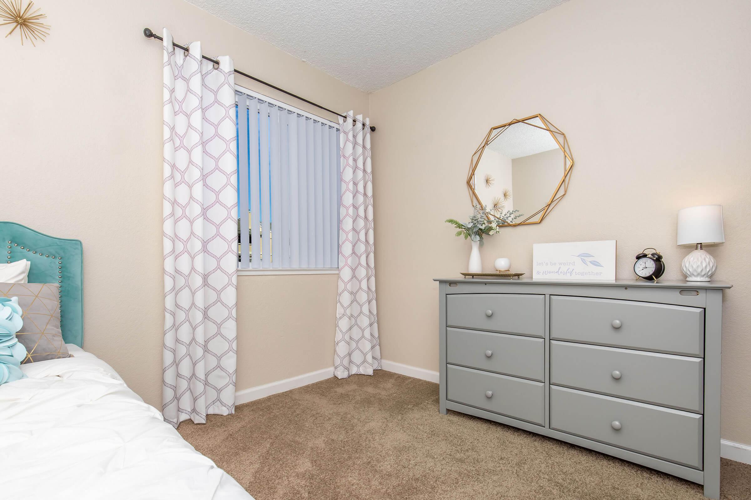 A cozy bedroom featuring a gray dresser, a large round mirror, light-colored walls, and decorative curtains with a geometric pattern. A neatly made bed with blue and white pillows is on the left side, while a small table lamp sits on the dresser. Natural light is coming through a window with blinds.