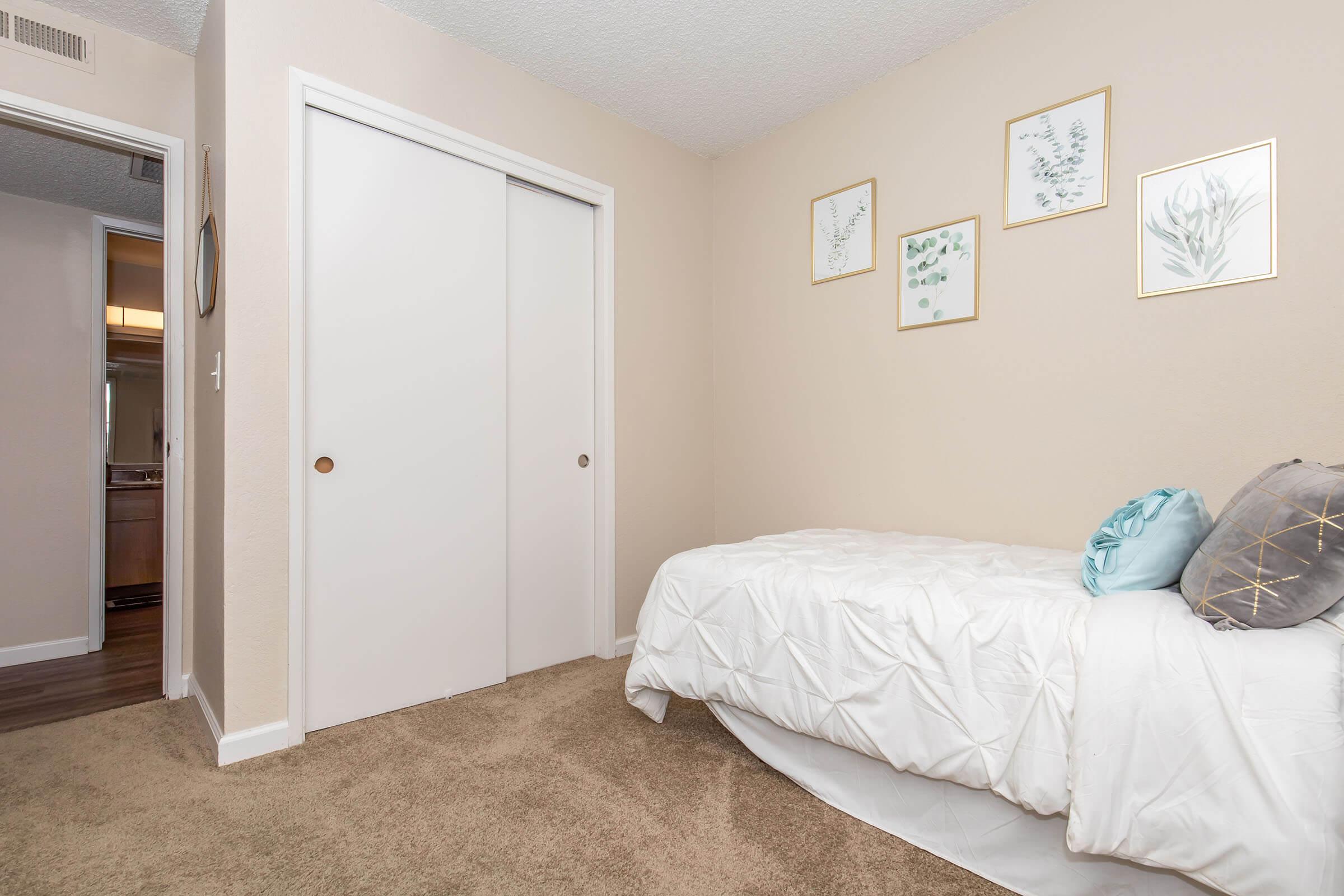 A simple bedroom featuring a neatly made bed with a white quilt, a decorative pillow, and light beige walls. There are framed botanical prints mounted on the wall above the bed and a closet with sliding doors. The floor is covered in a soft, light brown carpet.