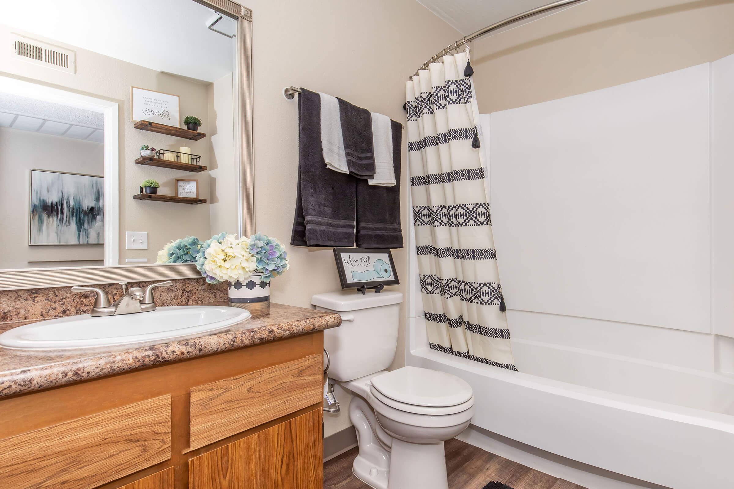 A well-lit bathroom featuring a light-colored countertop, a sink with a modern faucet, a white shower curtain with a geometric pattern, a bathtub, and a toilet. A small shelf holds decorative items, and blue and white hydrangeas are displayed on the counter. Neutral walls create a calm atmosphere.