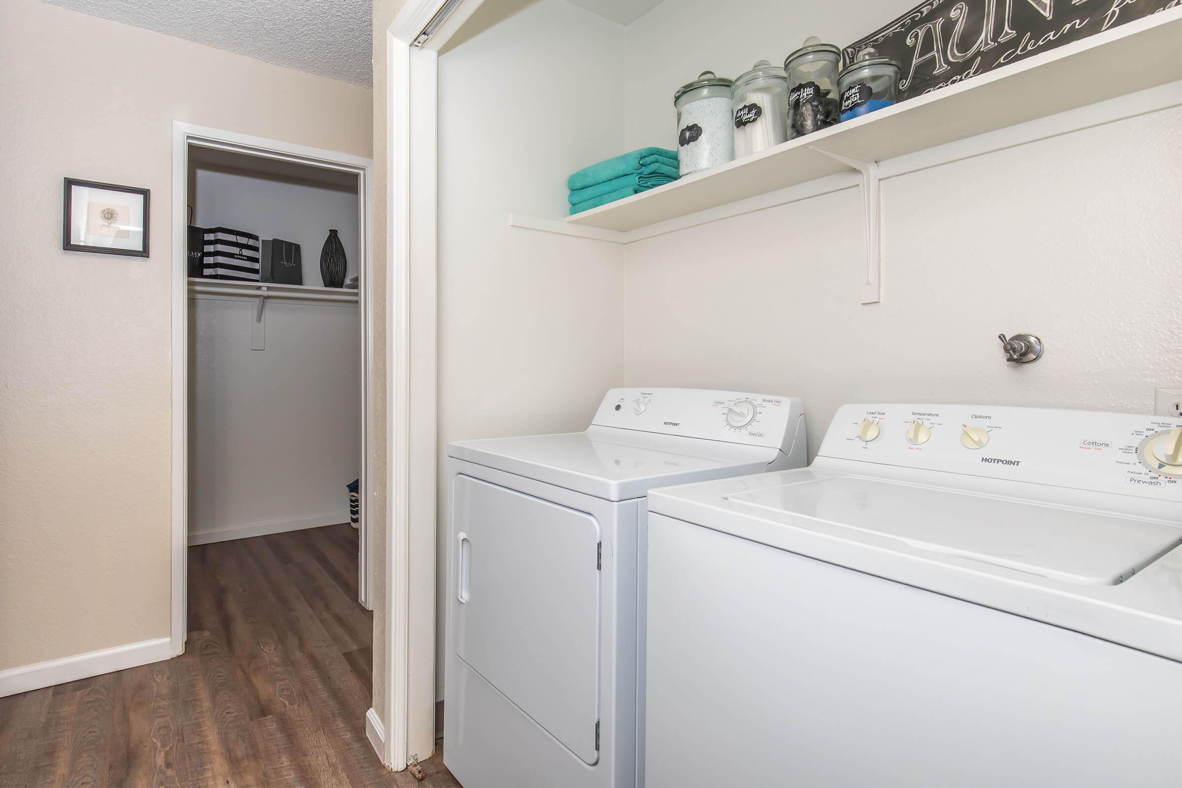 A laundry room featuring a white washer and dryer side by side. Above the appliances are shelves with jars, towels, and other items neatly organized. To the left, there's a doorway leading to a closet, with a simple framed picture on the wall. The floor has a modern wood-like finish.