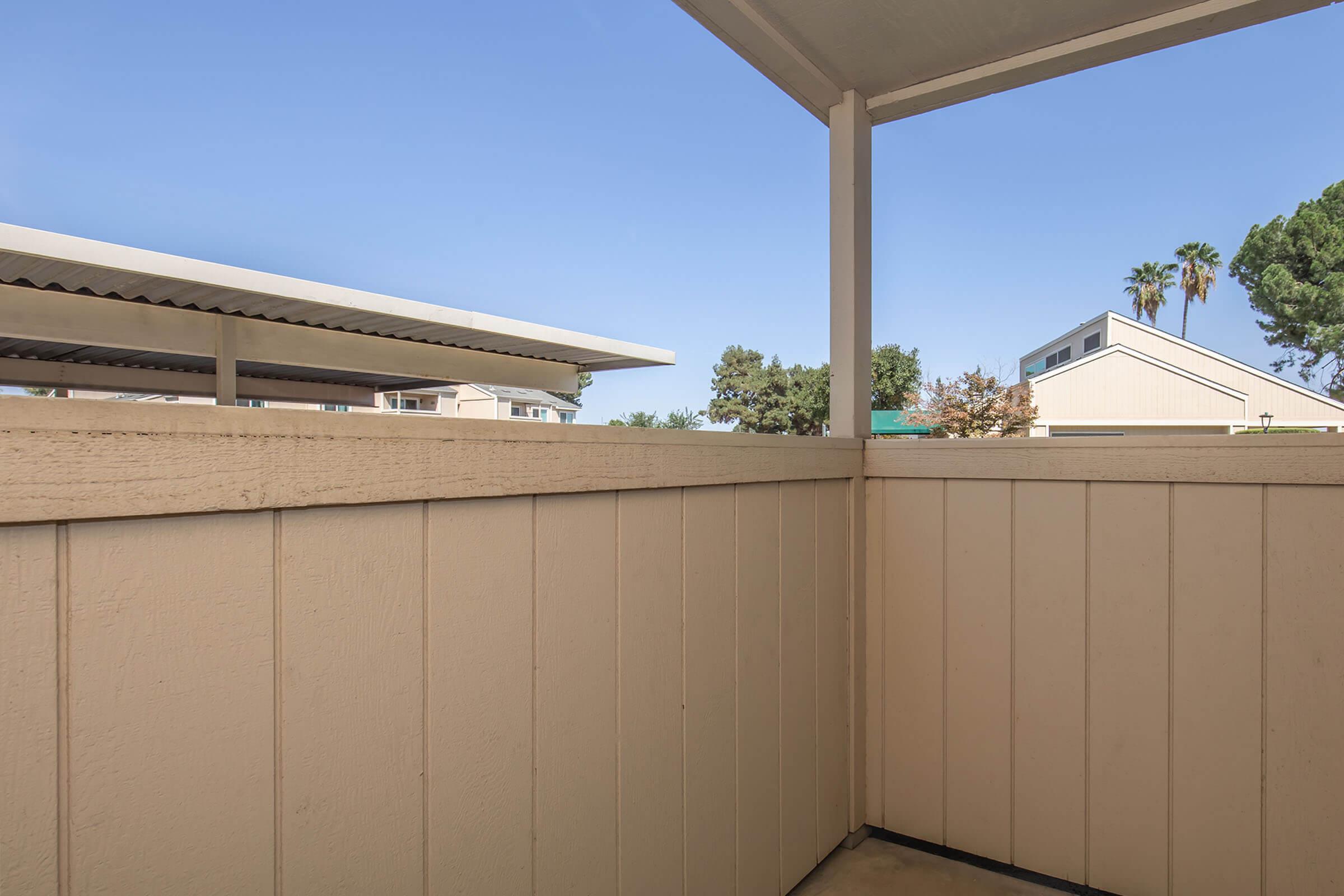 A view from a balcony featuring a wooden railing and wall. In the background, there are some residential buildings and palm trees under a clear blue sky. The setting suggests a sunny day in a suburban area.