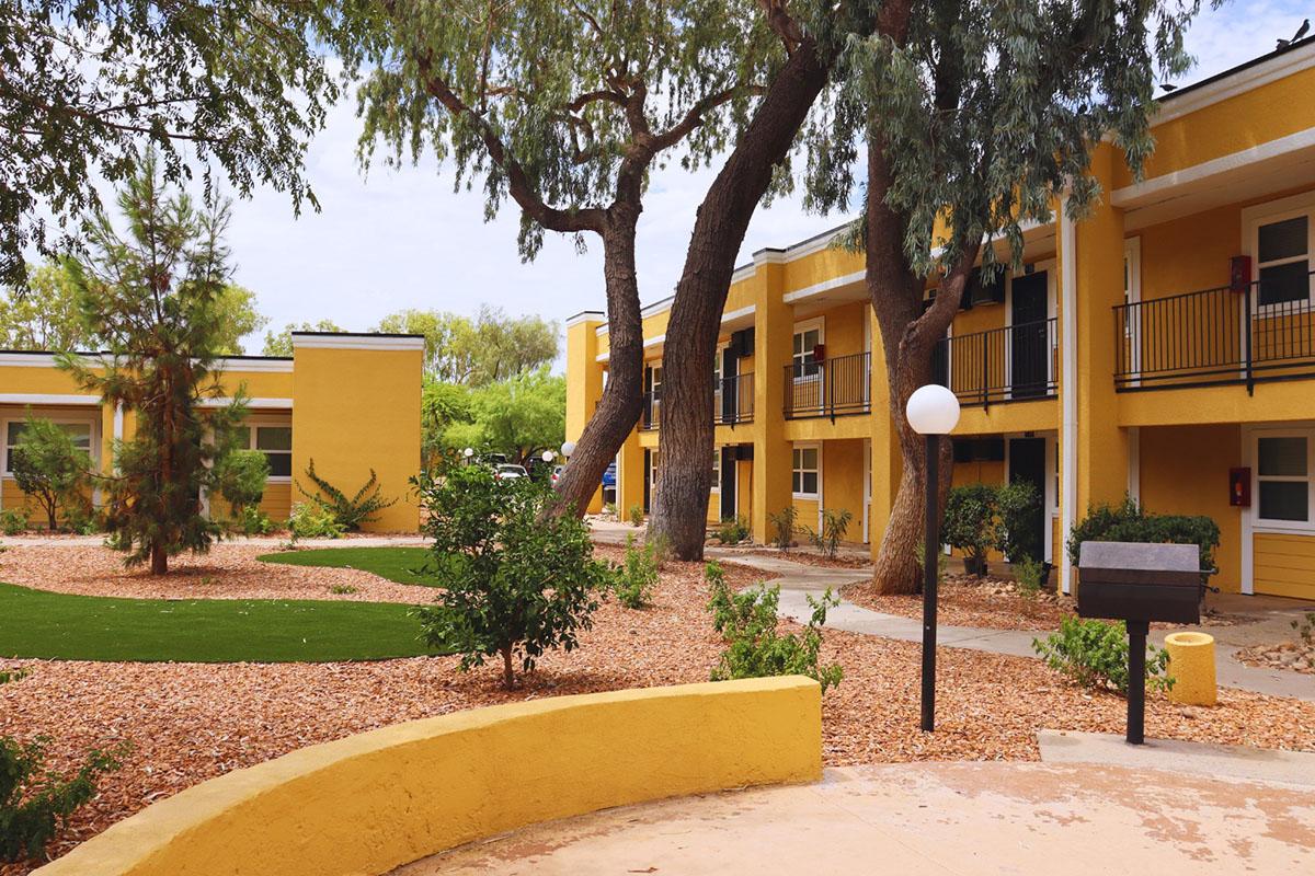 A courtyard of a yellow building complex featuring several trees and shrubs. The area has a well-maintained lawn and decorative rock landscaping. There are balconies overlooking the courtyard, creating a welcoming atmosphere. A pathway leads through the space, with a mailbox and light fixture visible.