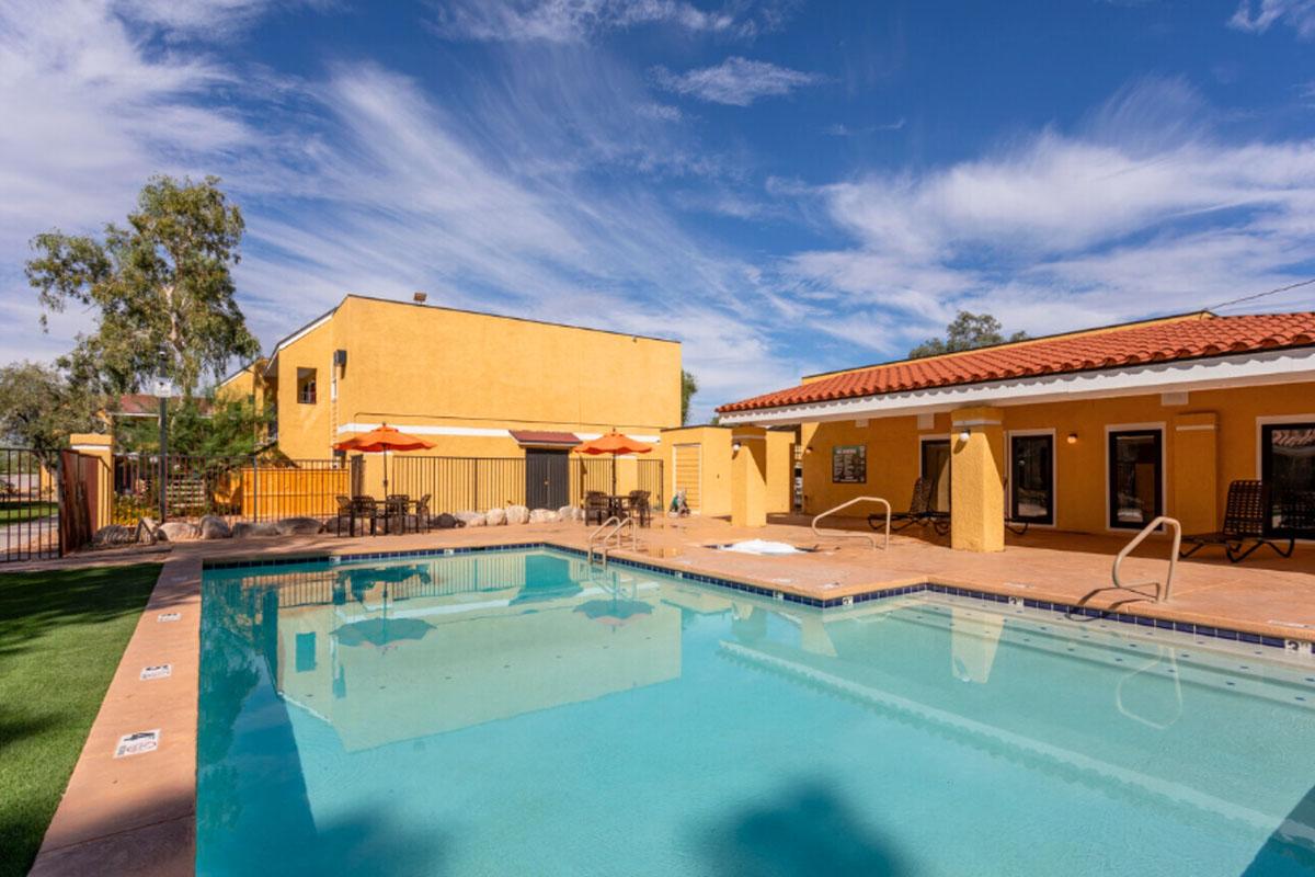A sunny outdoor pool area with a clear blue water, surrounded by lounge chairs and orange umbrellas. The backdrop features a yellow building, trees, and a clear blue sky with white clouds. The setting provides a relaxing and inviting atmosphere for leisure activities.