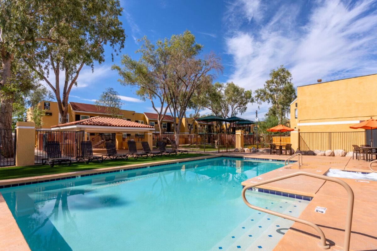 A clear blue swimming pool surrounded by lounge chairs and shaded areas, set in a landscaped outdoor area with trees. In the background, there are residential buildings and umbrellas providing shade. The sky is bright and sunny, creating a relaxing atmosphere.