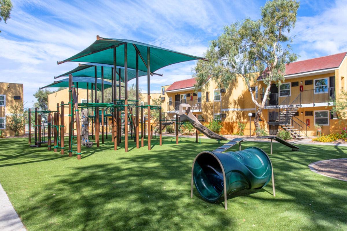 A playground with green turf featuring climbing structures, slides, and tunnels, surrounded by trees and adjacent to residential buildings. The scene is bright and inviting under a blue sky with scattered clouds.