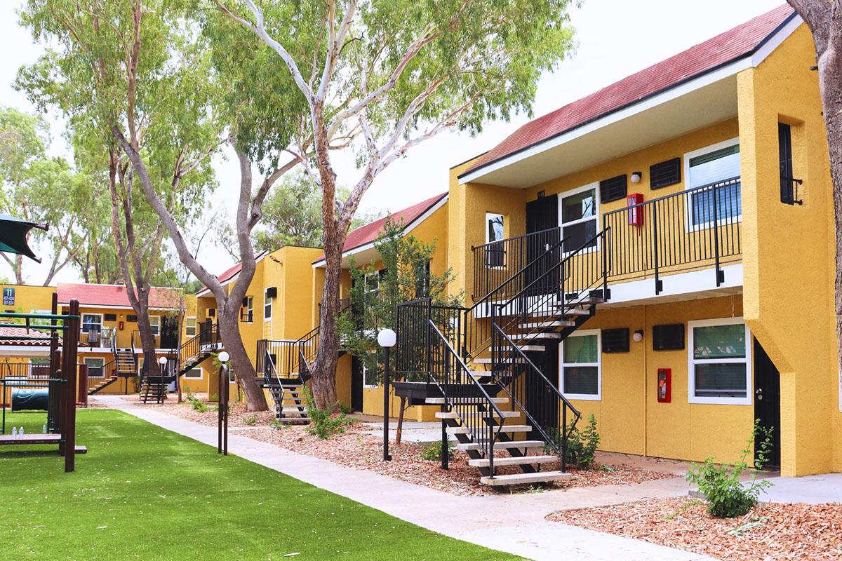 A courtyard featuring several yellow apartment buildings with balconies, surrounded by trees. There is a grassy area in the foreground and a playground structure visible. The buildings have black railings and various windows, creating a bright and inviting atmosphere.