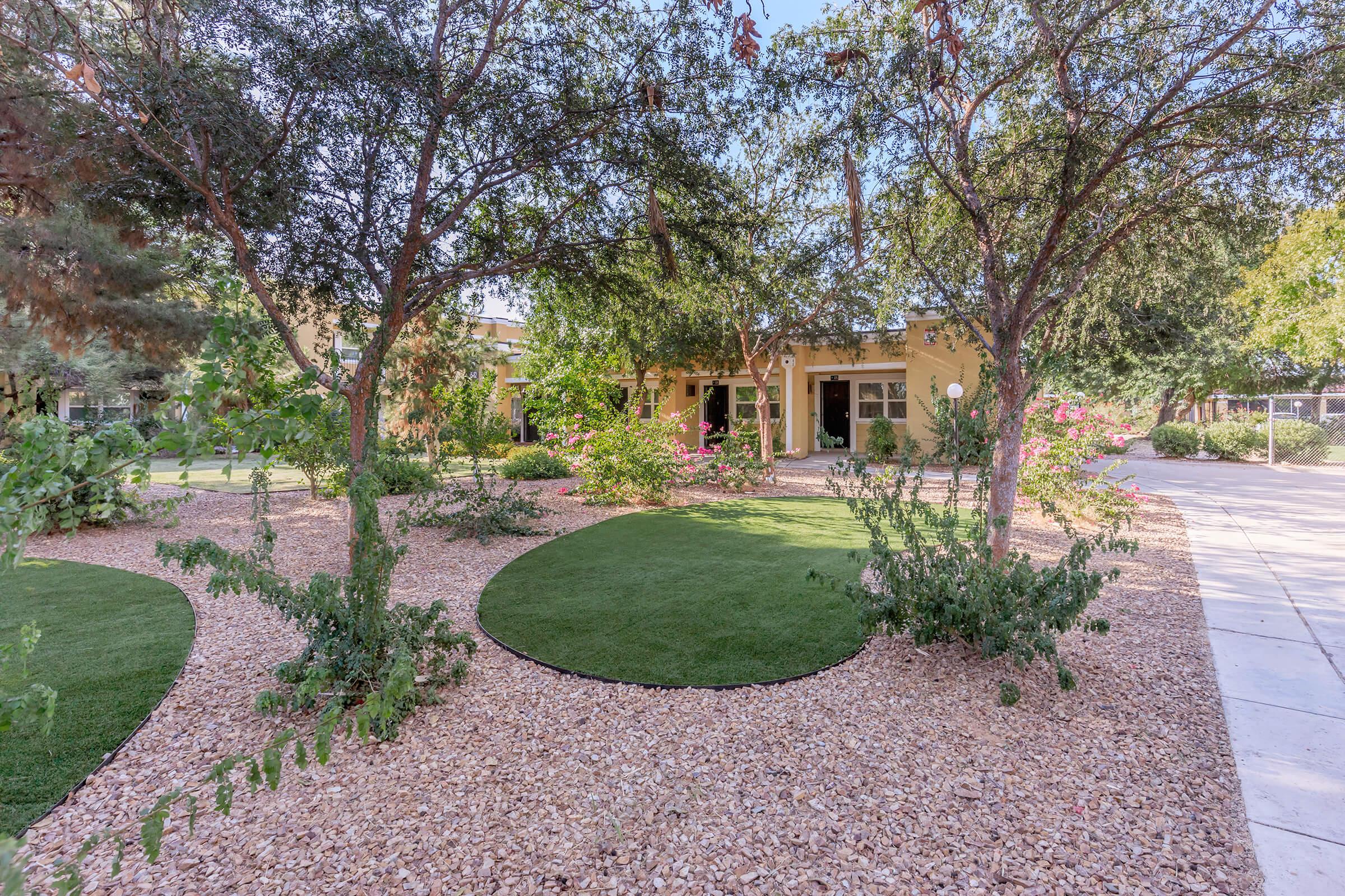 A landscaped garden featuring circular patches of green grass surrounded by decorative stones. Lush greenery, including trees and colorful flowers, creates a vibrant outdoor space. A building with open entrances is visible in the background, adding to the serene atmosphere of the setting.