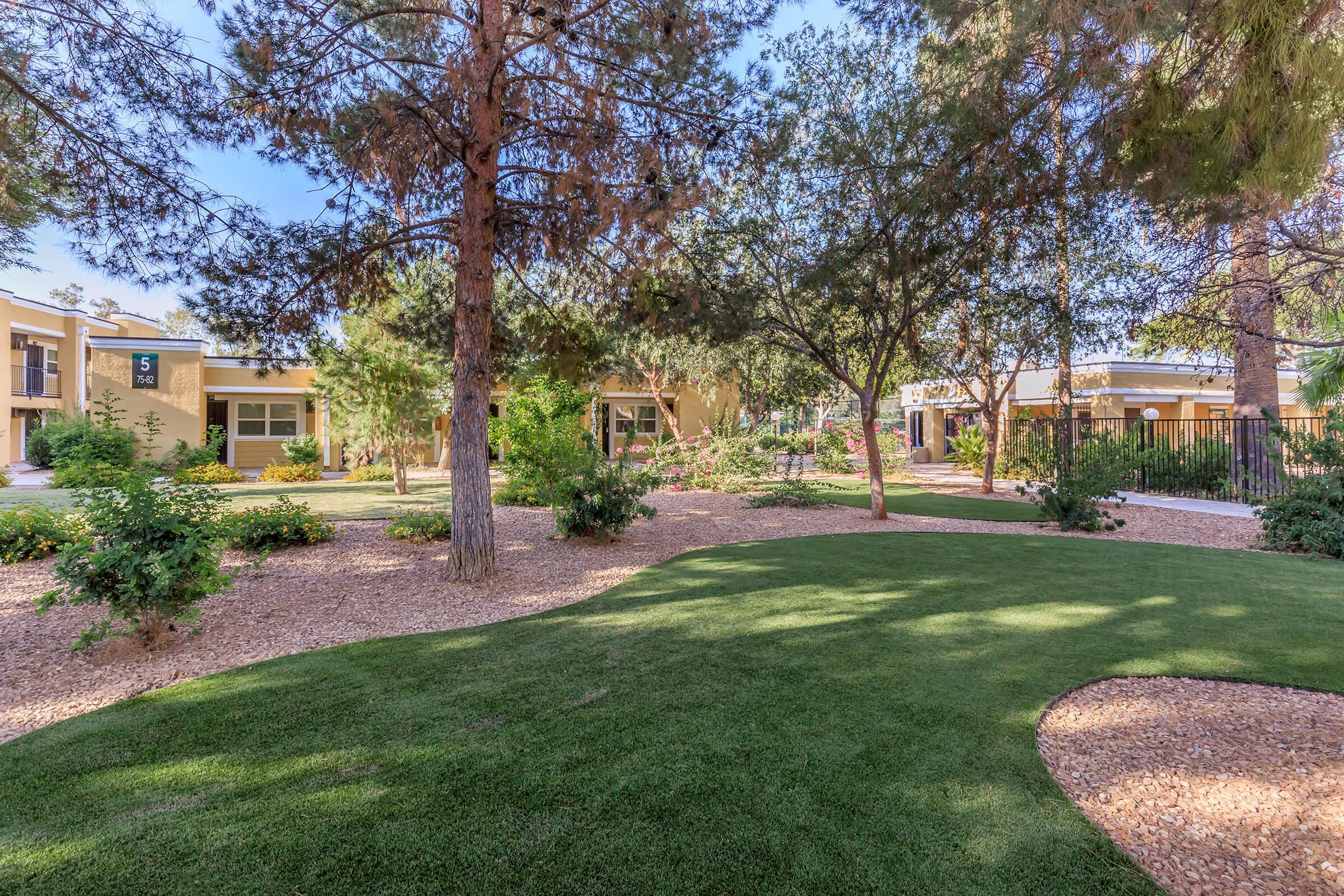 A landscaped garden featuring well-maintained grassy areas surrounded by trees and shrubs. Pathways meander through the space, leading to buildings with patios. The scene is illuminated by clear blue skies, creating a serene and inviting atmosphere.